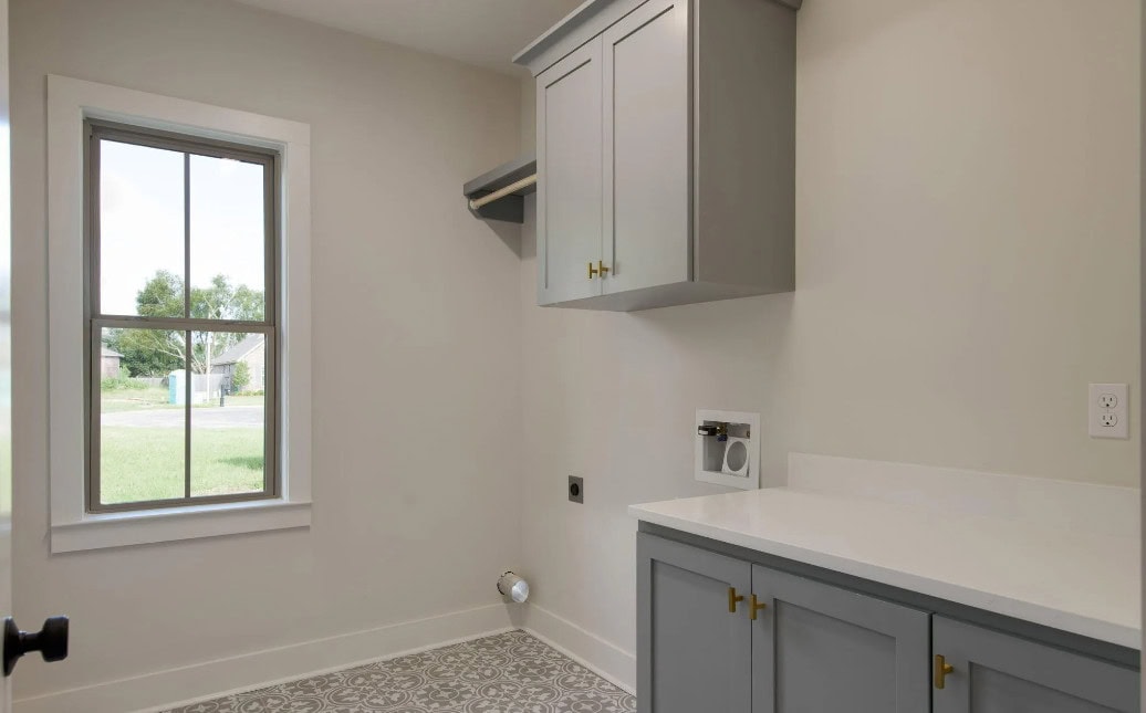 A laundry room with light gray cabinets, a white countertop, patterned tile flooring, and a single window providing natural light. There are wall outlets and plumbing for a washer and dryer.