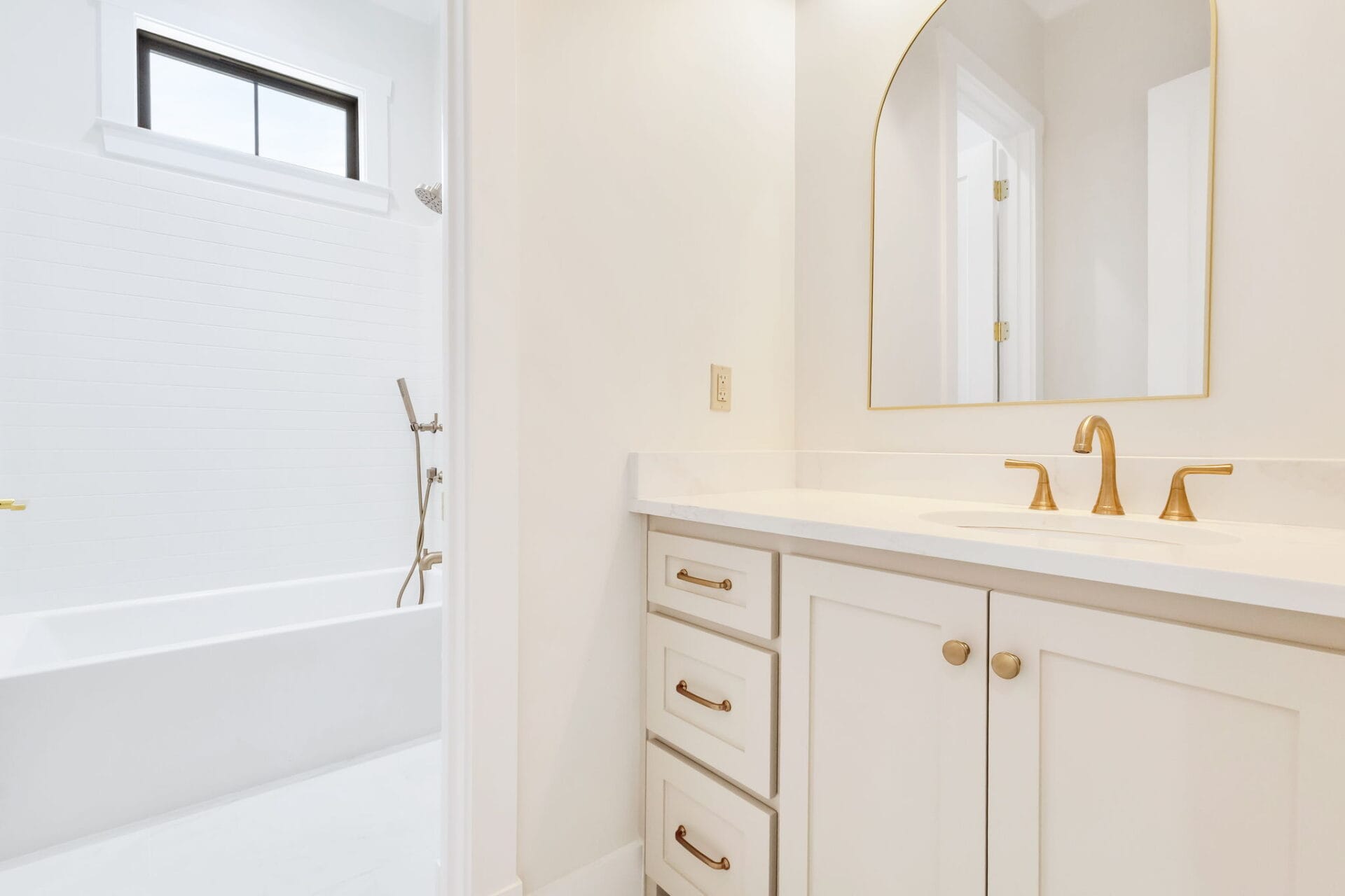 A bathroom with white cabinets and gold hardware, a white countertop with a gold faucet, an arched mirror, and a white bathtub with a gold shower fixture. Walls are white and the room is well-lit.