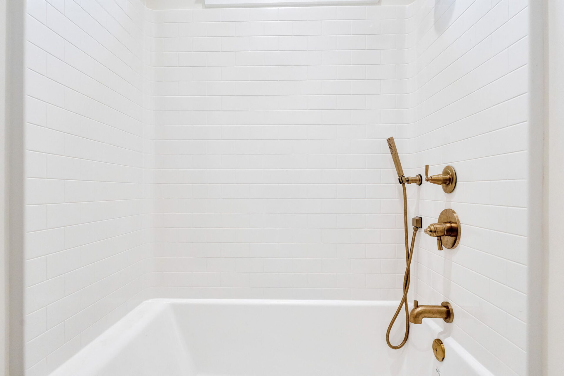White bathtub and shower combination with white subway tile walls, featuring gold fixtures including a handheld showerhead, faucet, and two control knobs. The setting is clean and minimalistic.