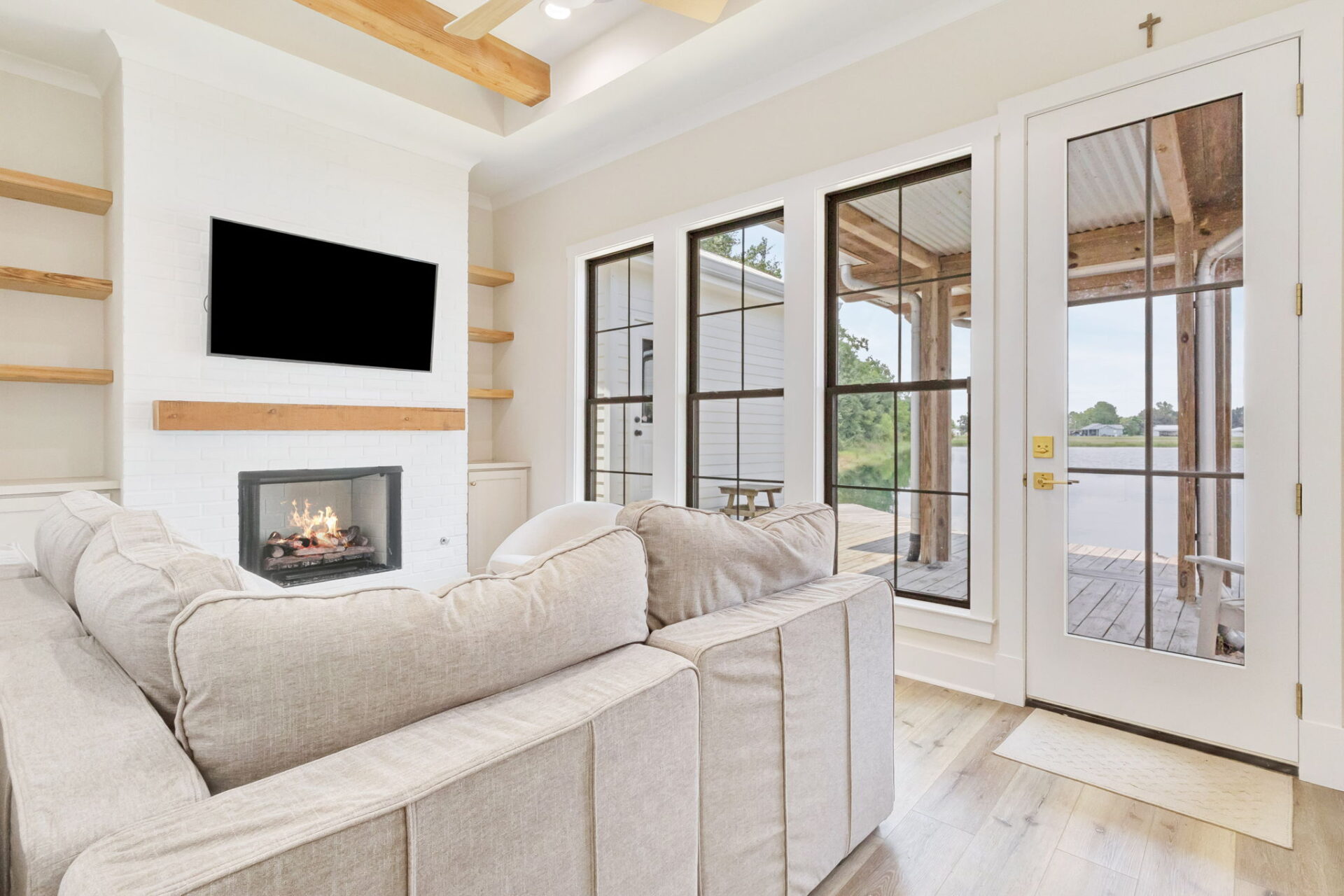 A bright living room with a beige sectional sofa, a fireplace with a wooden mantel, a wall-mounted TV, wooden shelves, large windows, and a glass door leading to a covered deck overlooking water.