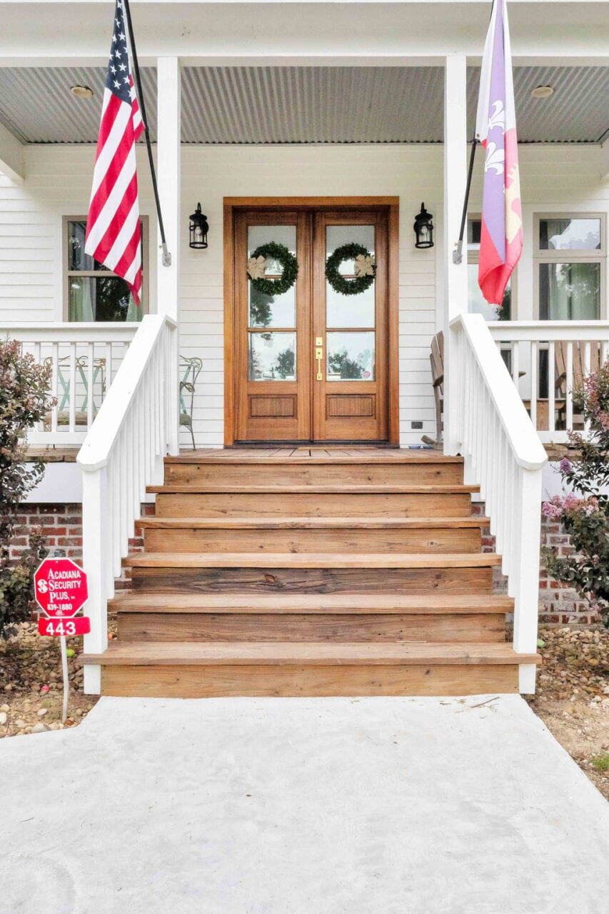 A front porch with wooden steps leading up to double doors decorated with green wreaths. Two flags are mounted on either side, and a red security sign is displayed near the bottom left of the steps.