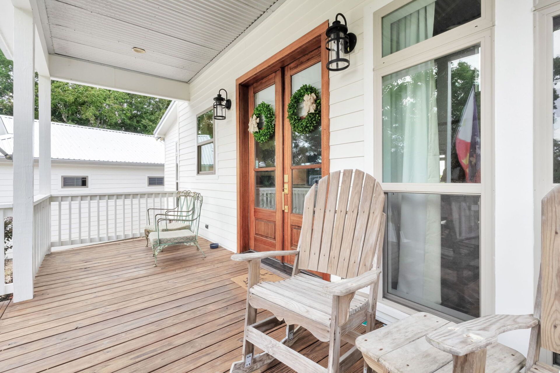 A wooden front porch with two Adirondack chairs, a metal chair, double wooden doors with wreaths, a window with curtains, white siding, and outdoor wall lanterns, with part of another house visible in the background.