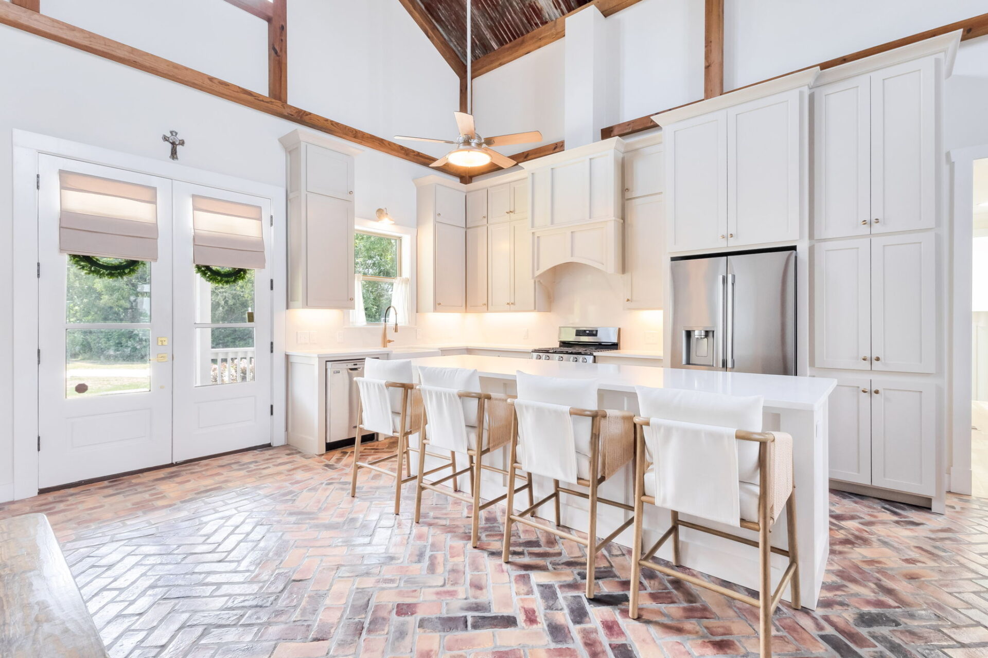 A bright kitchen with white cabinets, stainless steel appliances, a large island with four wood and white bar stools, brick herringbone floor, ceiling fan, and double doors with wreaths.
