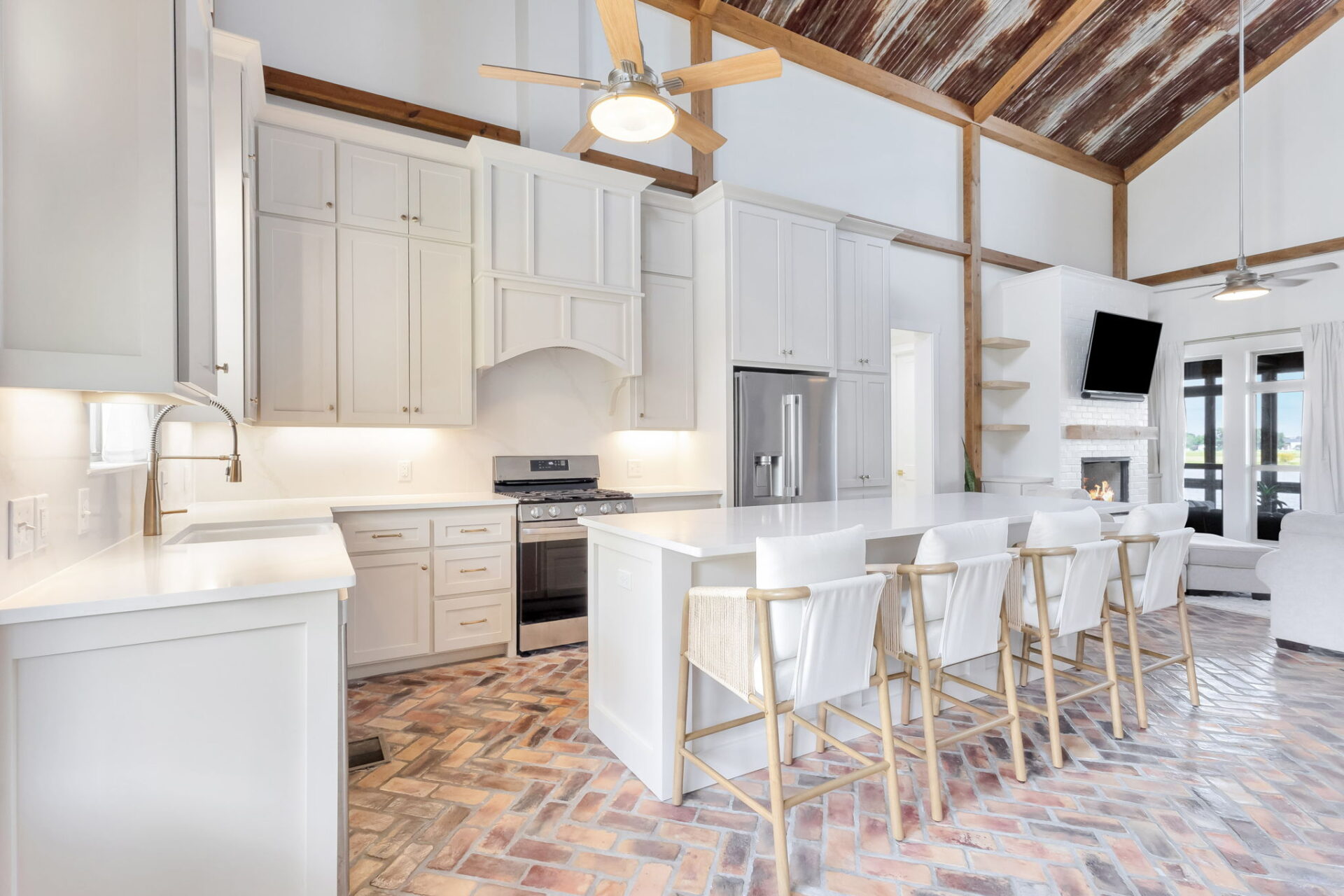 A modern kitchen with white cabinets, stainless steel appliances, a large island with five white barstools, brick herringbone flooring, exposed wood ceiling, and a ceiling fan.
