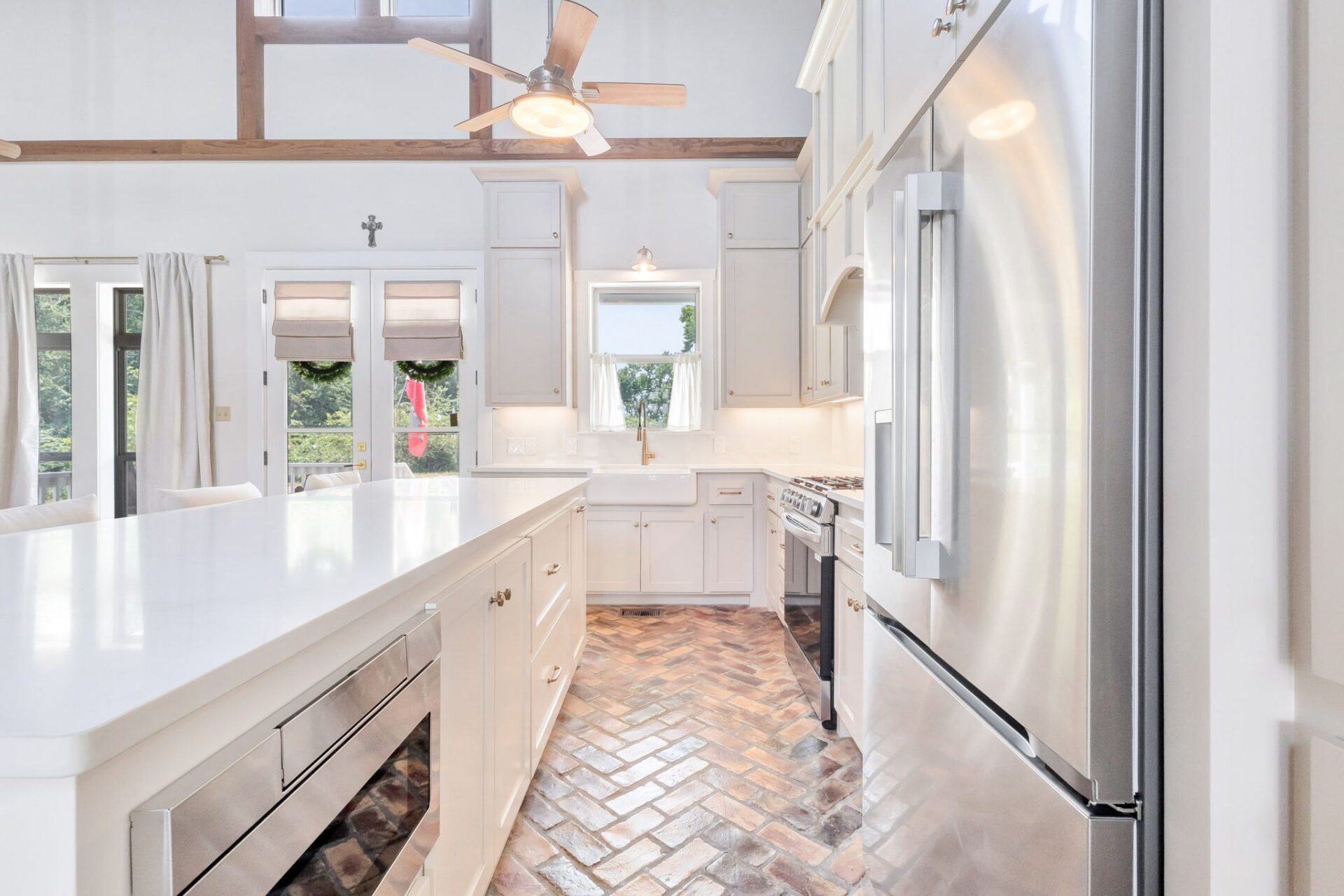 Bright, modern kitchen with white cabinets, stainless steel appliances, a large island, and herringbone-pattern brick flooring. Large windows and glass doors let in natural light, creating an open and airy atmosphere.