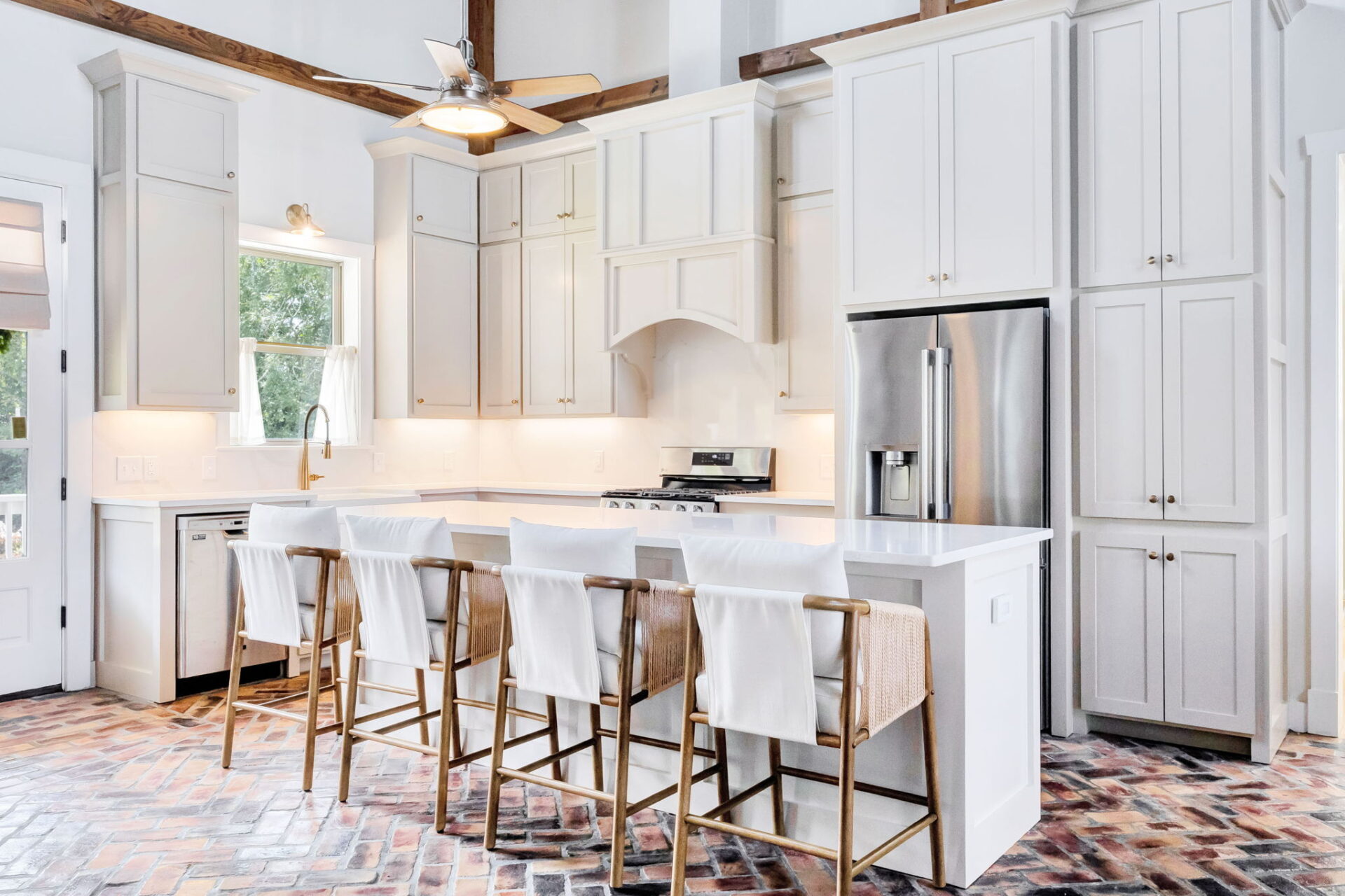 A modern kitchen with white cabinets, stainless steel appliances, and a large island with four barstools. The floor is brick-patterned, and the space has exposed wooden ceiling beams and a ceiling fan.