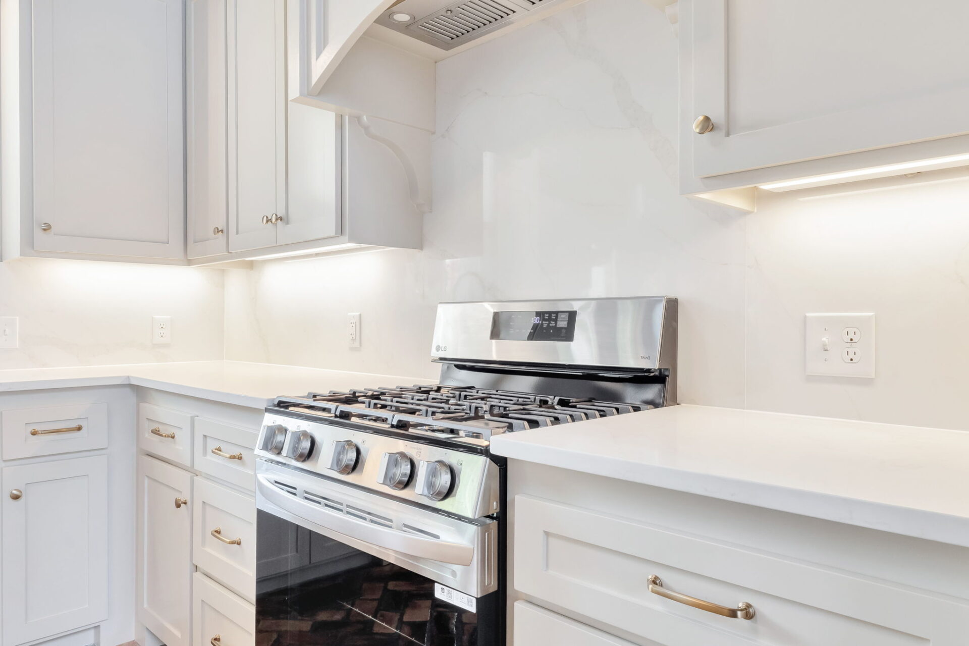 A modern kitchen with white cabinets, a stainless steel gas stove and oven, marble backsplash, white countertops, and under-cabinet lighting.