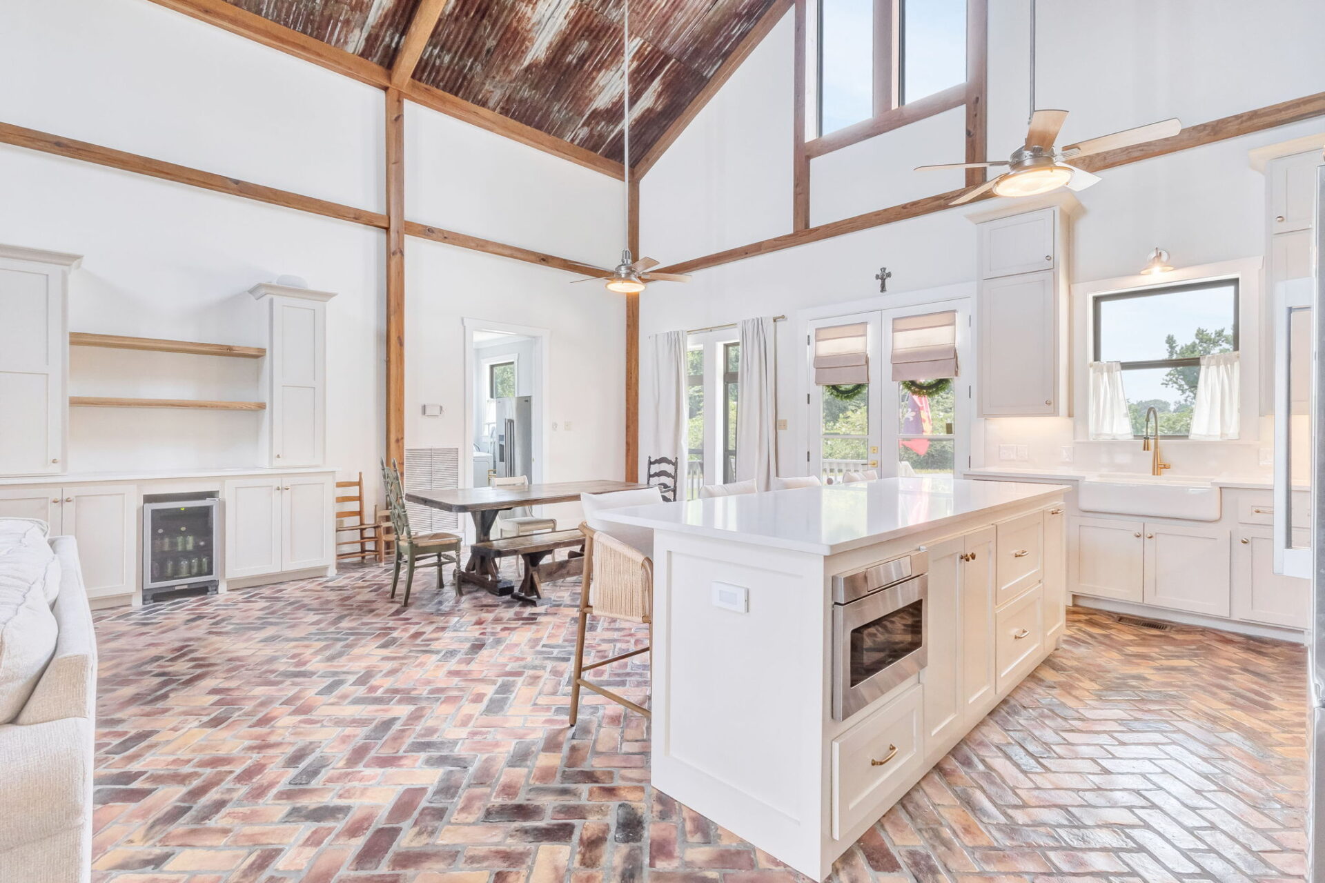 A spacious kitchen with high ceilings, exposed wooden beams, white cabinets, a large island with built-in appliances, a farmhouse sink, and brick-patterned tile flooring. Large windows let in natural light.
