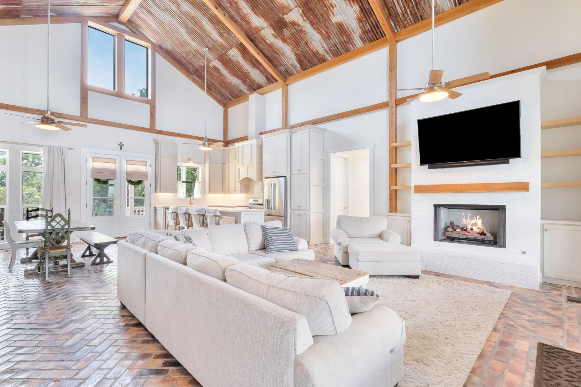 Spacious open-concept living room with vaulted, rustic wood ceiling, white walls, brick floor, modern kitchen, large beige sectional sofa, fireplace, mounted TV, and tall windows letting in natural light.
