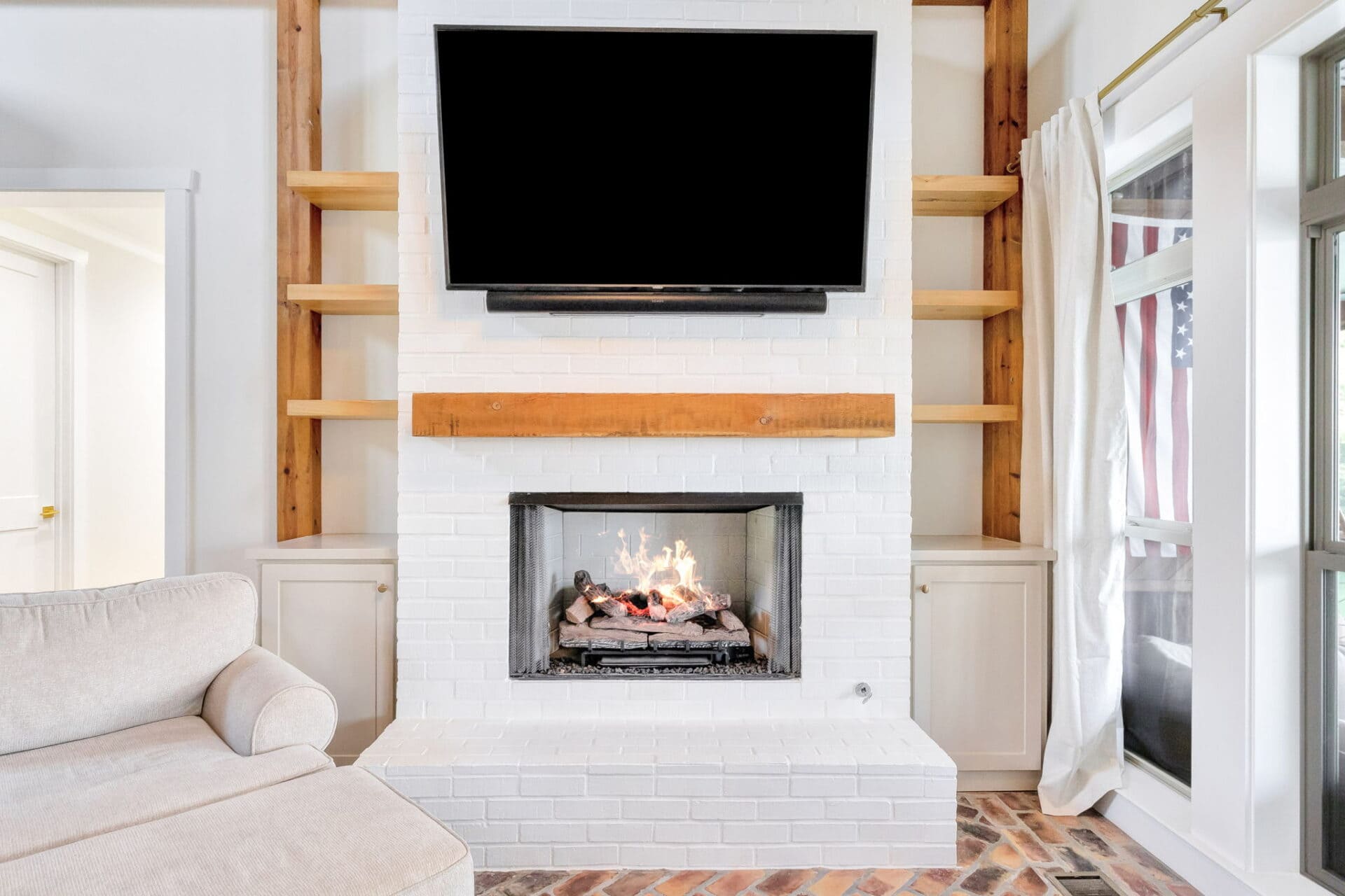 A modern living room features a white brick fireplace with a wooden mantel, a wall-mounted flat-screen TV above it, built-in shelves on each side, and a beige sectional sofa in the foreground.