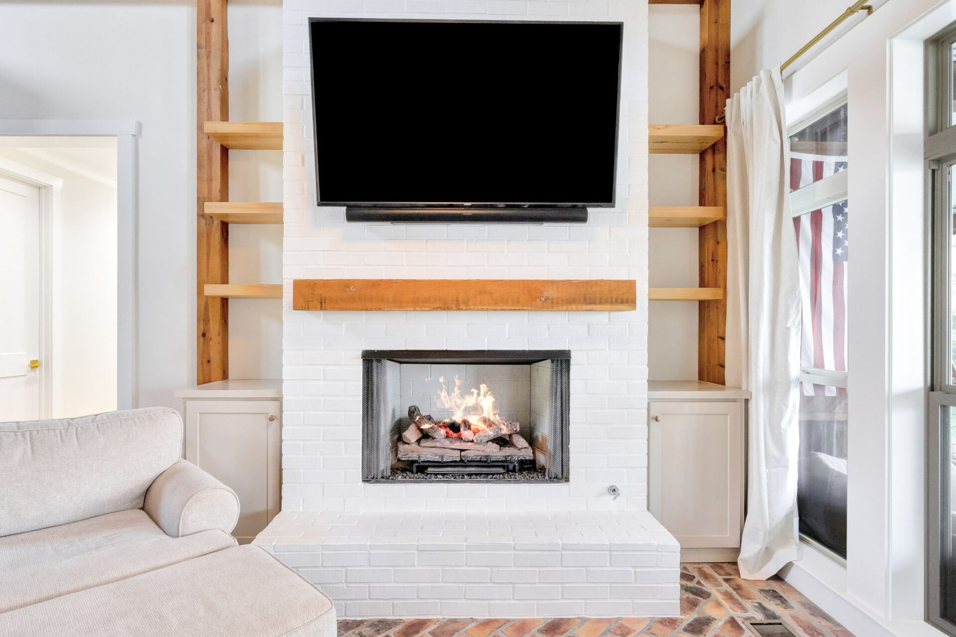 A white brick fireplace with a wooden mantel, a lit fire inside, and a large flat-screen TV mounted above. Built-in shelves and cabinets are on either side, with part of a beige sofa in the foreground.