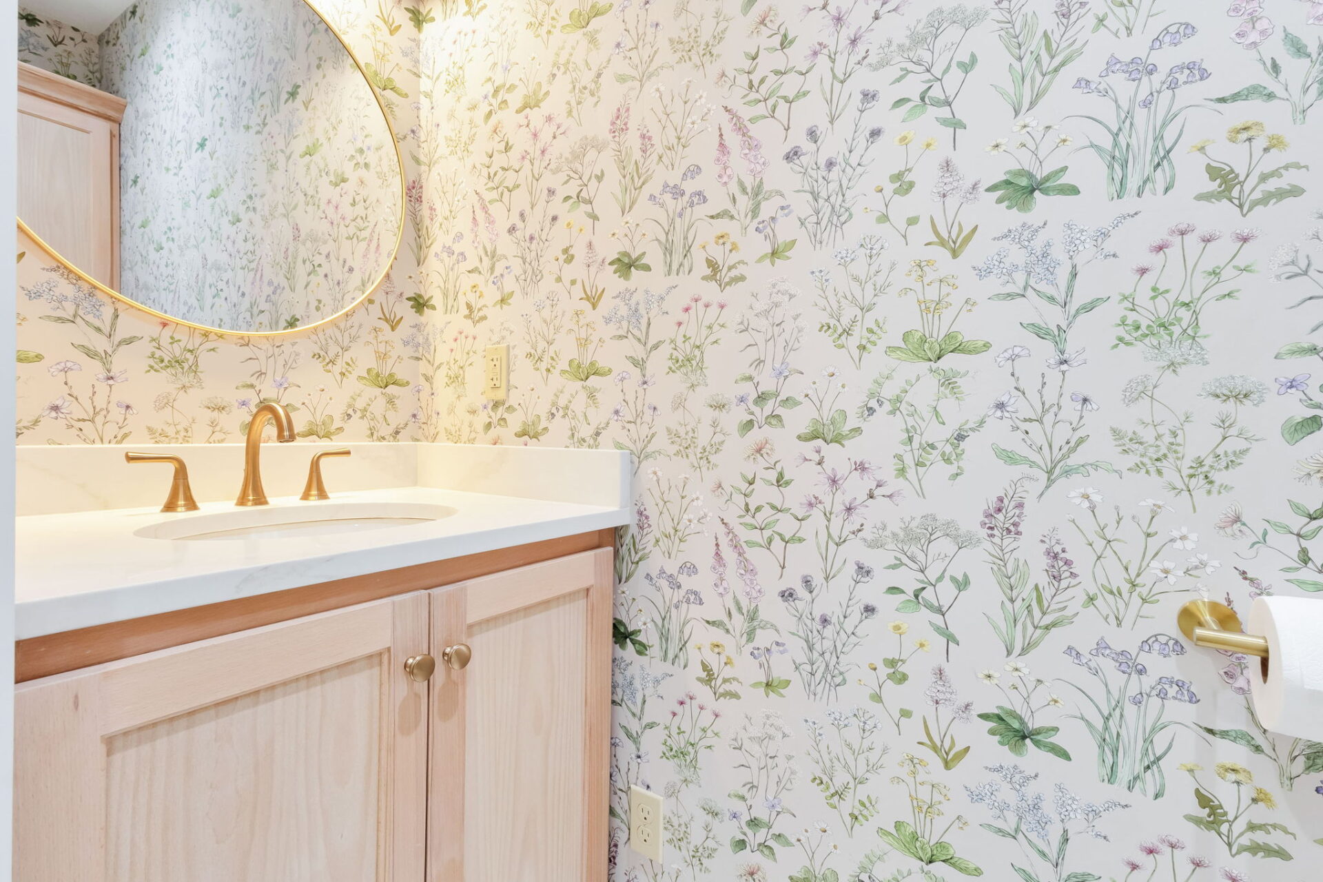 A bathroom with a round mirror, a sink with gold fixtures, a marble countertop, light wood cabinets, and floral-patterned wallpaper. A toilet paper holder is visible on the wall to the right.