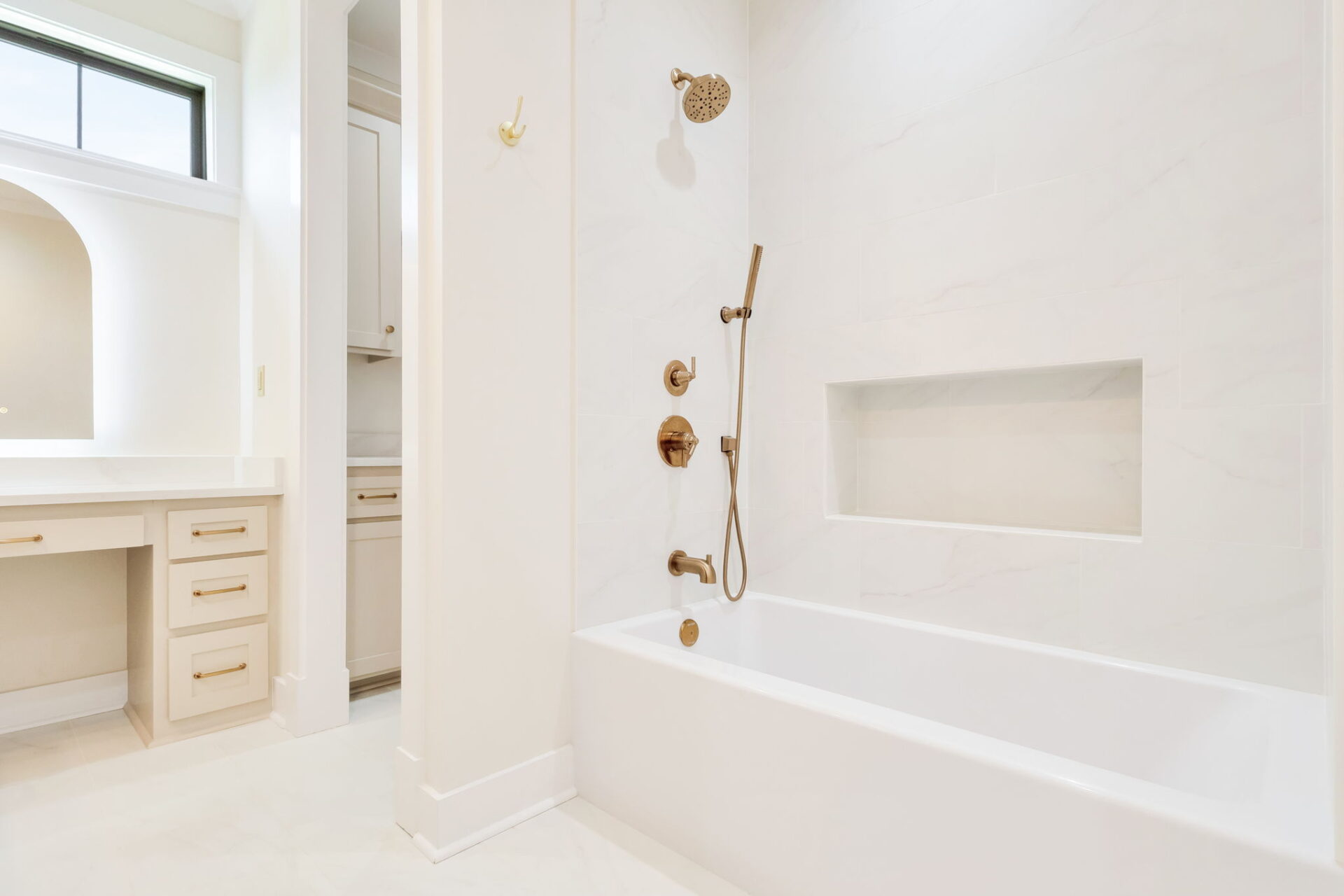 A modern bathroom with a white bathtub, built-in shower with gold fixtures, a recessed shelf in the shower wall, and a vanity with drawers and a mirror in the background. The room features light-colored walls and floors.