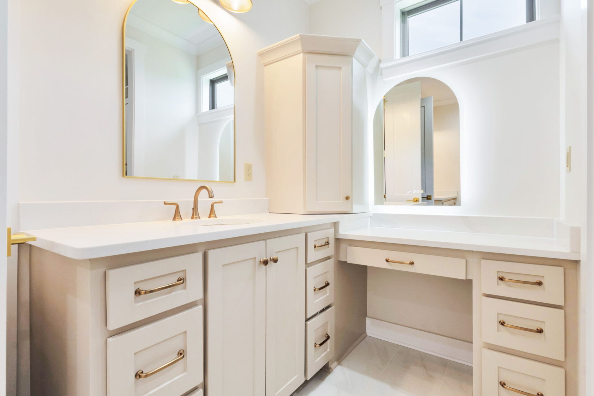 A modern bathroom features white cabinets with gold handles, a sink with a gold faucet, two mirrors with gold frames, and a built-in vanity area beneath one of the mirrors.