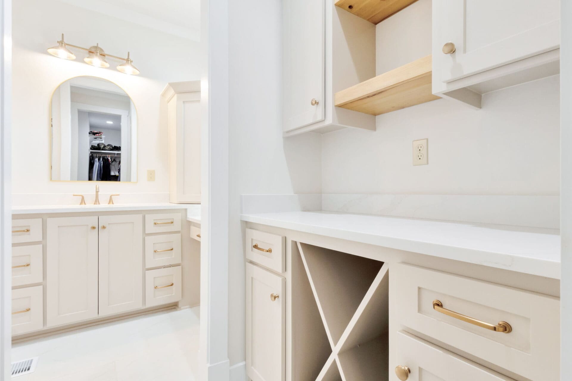 A bright, modern bathroom with cream cabinets, gold hardware, a double sink vanity with a large mirror, and a counter featuring open shelving and a wine rack-style storage area.
