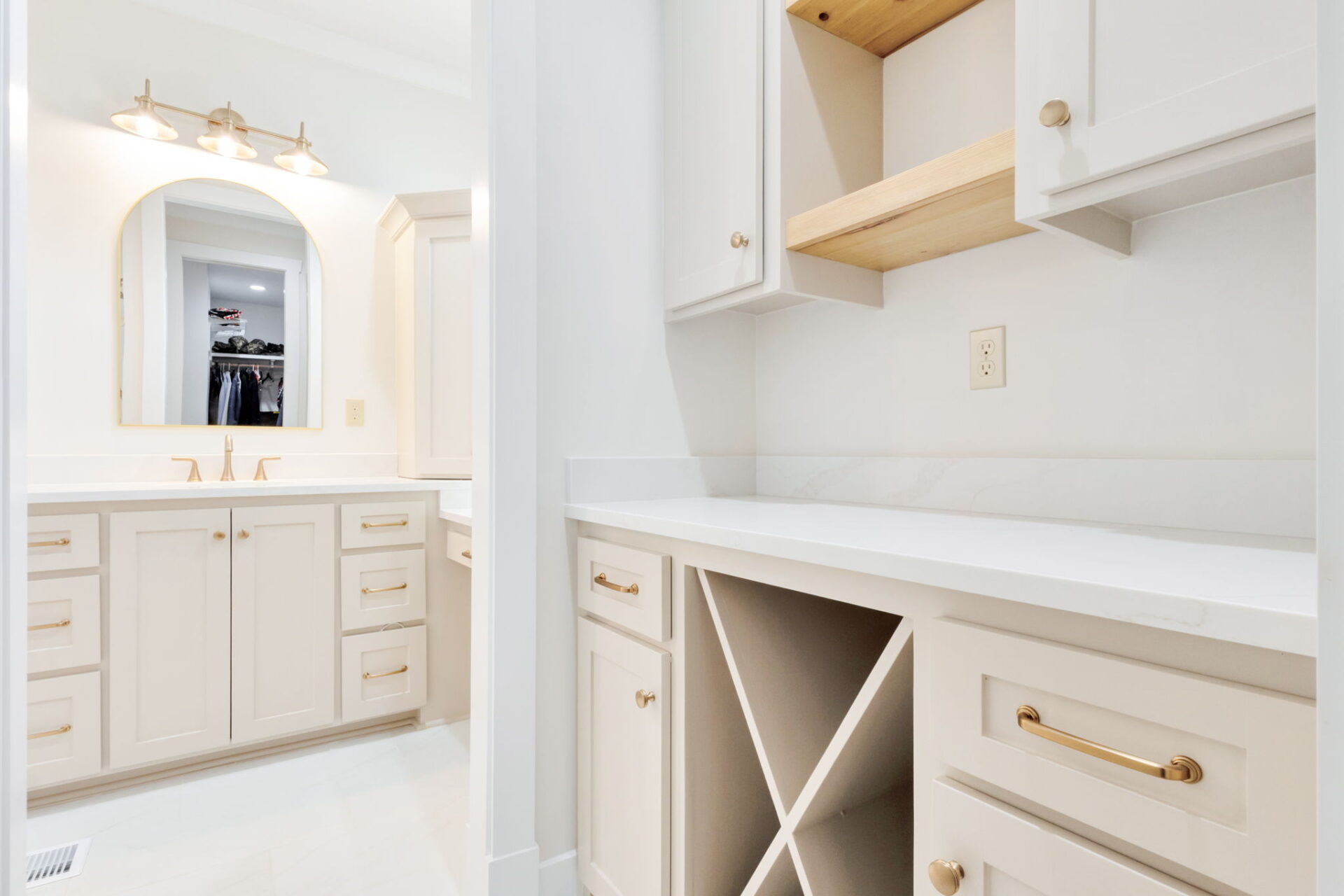 A bright bathroom with white cabinetry, gold hardware, an empty wine rack, and open shelving. A double sink vanity with a mirror and three-light fixture is visible in the background.