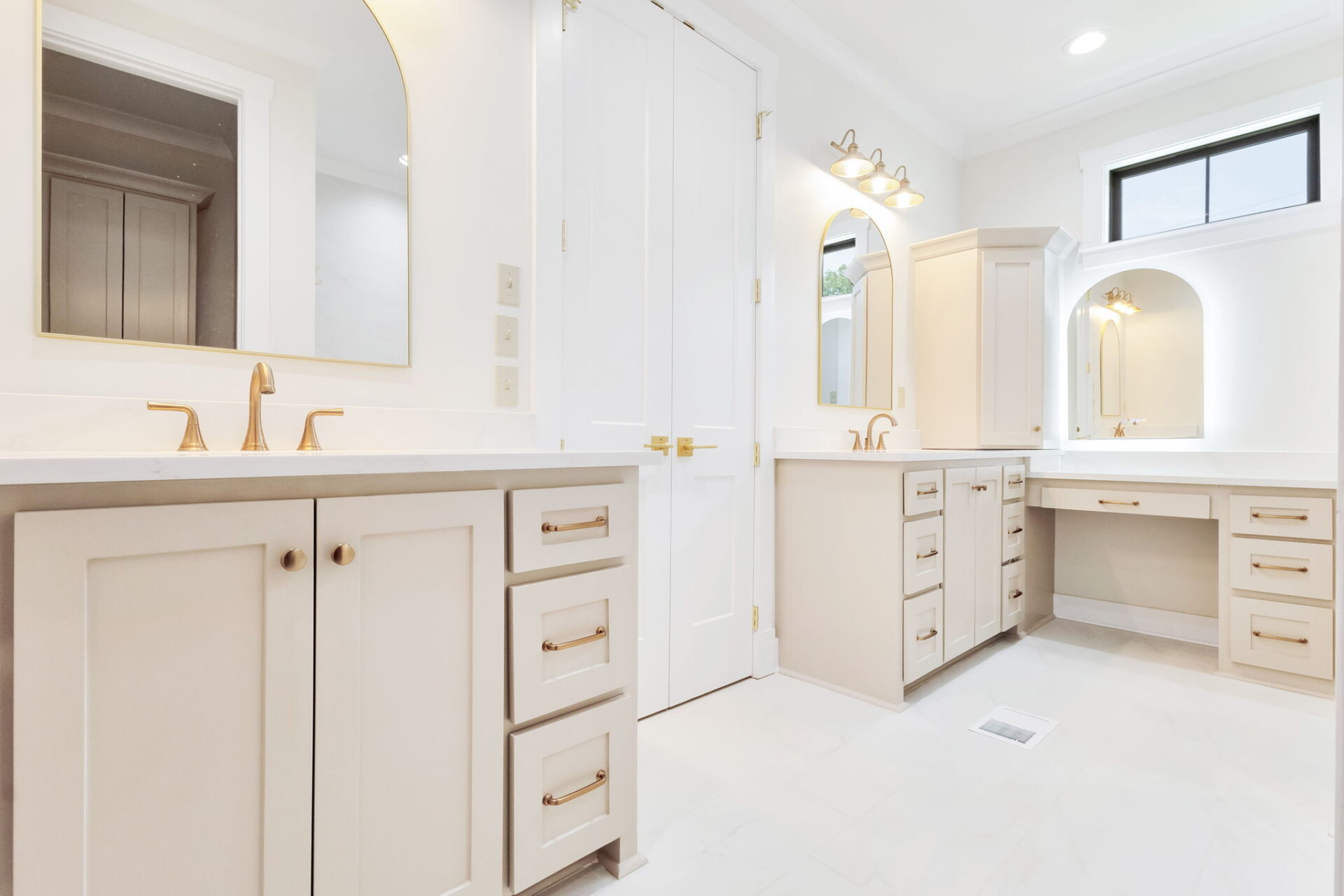 A spacious bathroom with beige cabinets, gold fixtures, two arched mirrors, a makeup vanity area, white countertops, and a double door in the center. The space is brightly lit with natural and artificial light.