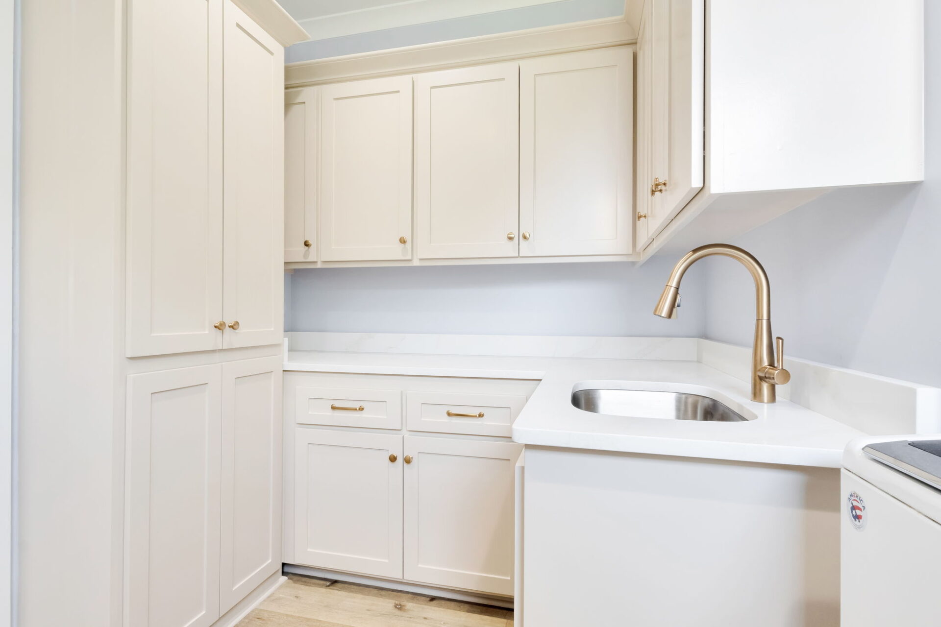 A modern kitchen with white cabinets, gold handles, a stainless steel sink with a gold faucet, white countertops, and light wood flooring. The cabinets have a simple, clean design.