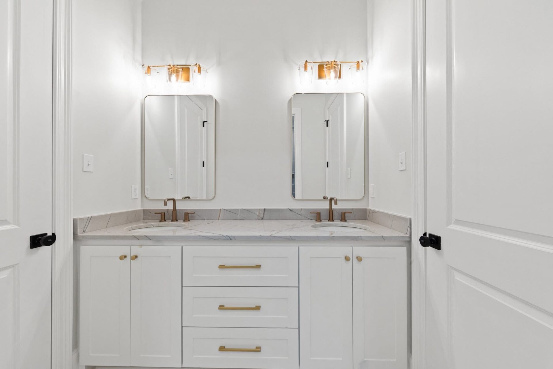 A white double-sink bathroom vanity with gold fixtures, two rectangular mirrors, and matching wall lights. The countertop is marble, and there are three drawers and two cabinets below. The walls and doors are white.