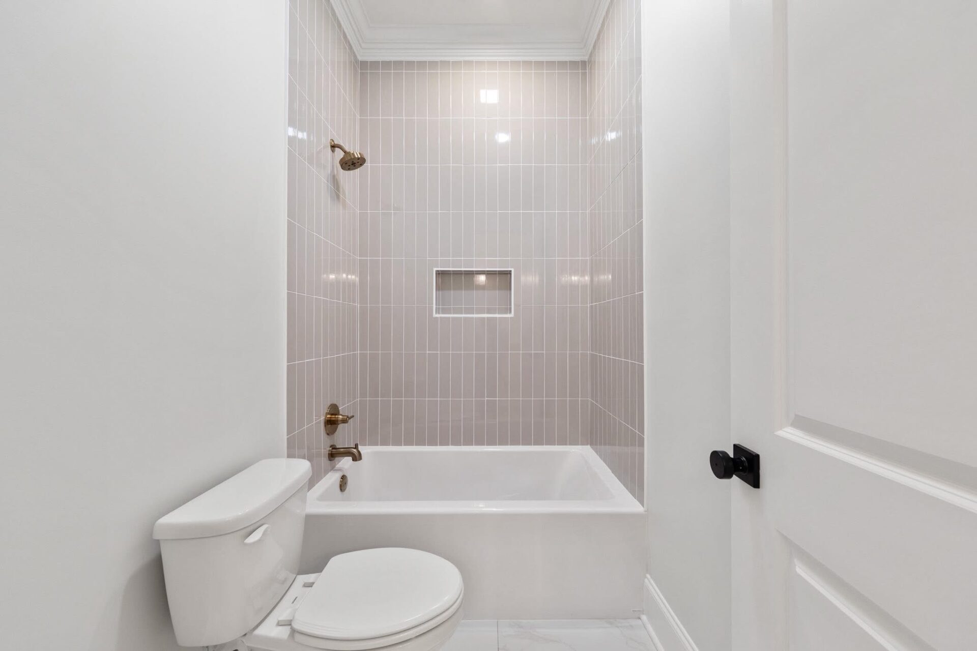 A white bathroom with a bathtub and shower, beige vertical wall tiles, a built-in niche, a white toilet, and a white door with a black handle. Fixtures are in a brass or gold finish.