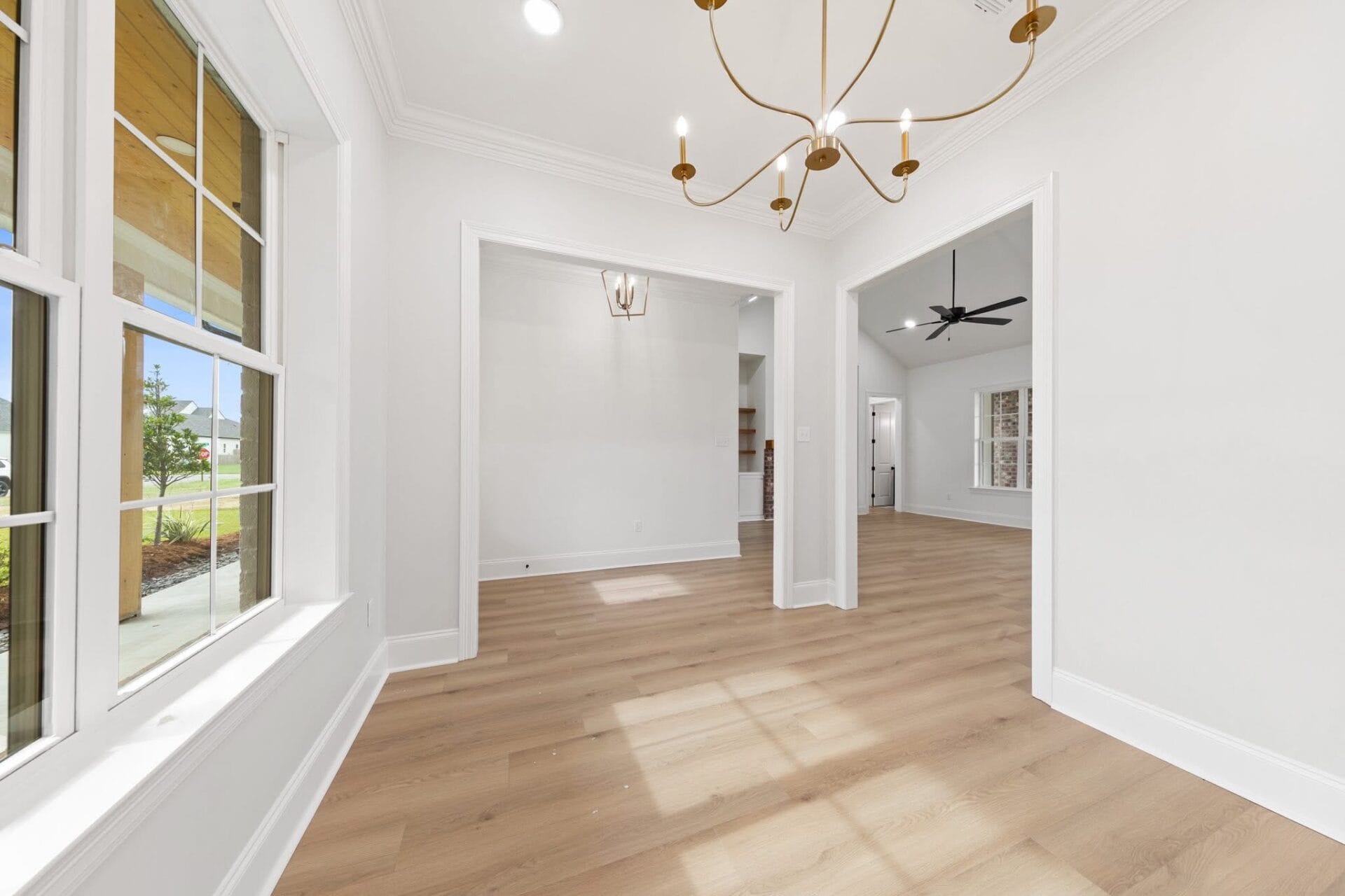 A bright, empty room in a modern house with light wood floors, large windows on the left, white walls, and a contemporary gold chandelier hanging from the ceiling. Two doorways lead to additional rooms.