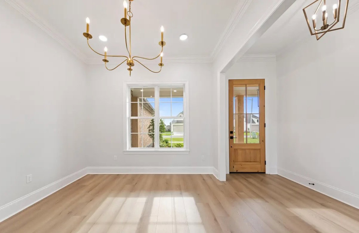 Bright, empty room with light wood floors, white walls, a large window, two modern gold chandeliers, and a wooden front door with glass panes letting in natural light.