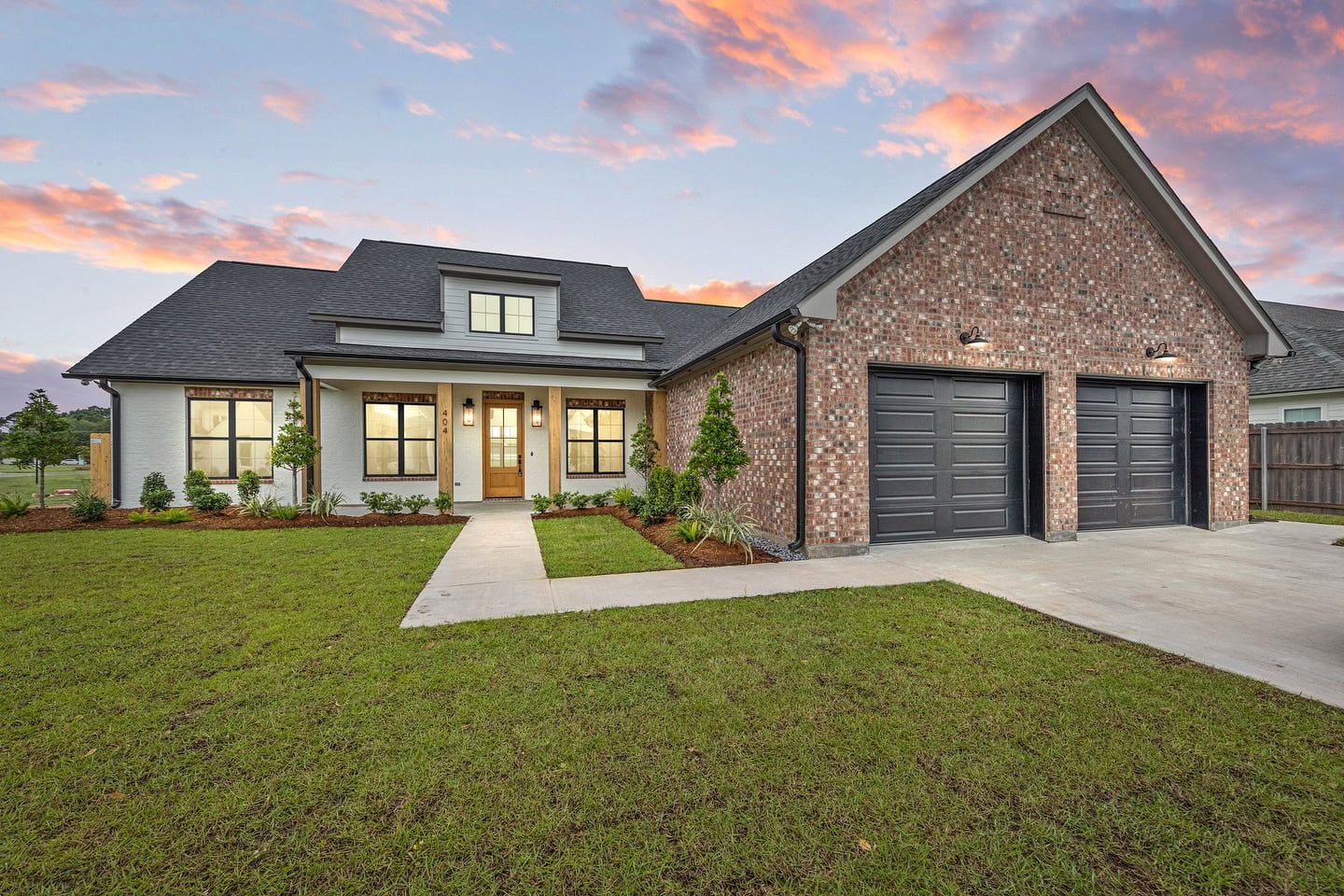 A modern single-story brick house with a covered front porch, double black garage doors, neatly landscaped yard, and a concrete driveway, set under a colorful sunset sky.