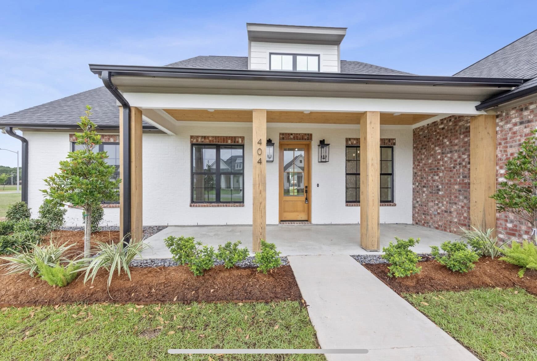 A one-story house with a covered front porch supported by wooden columns, white exterior walls, and brick accents. There are landscaped shrubs and plants along the walkway leading to a wooden front door.