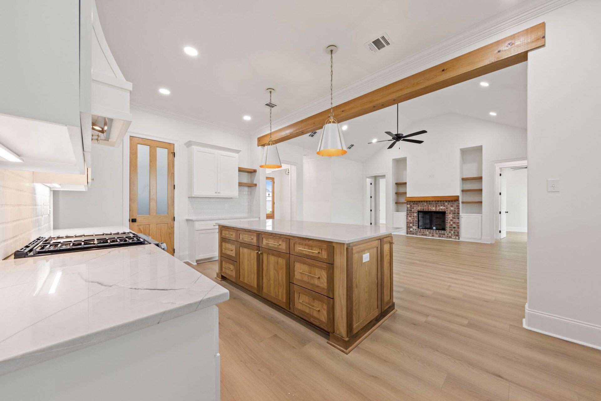 Modern kitchen with a large wooden island, white countertops, wood flooring, and pendant lights. The space opens to a living area with a brick fireplace, high ceiling, and exposed wooden beam.