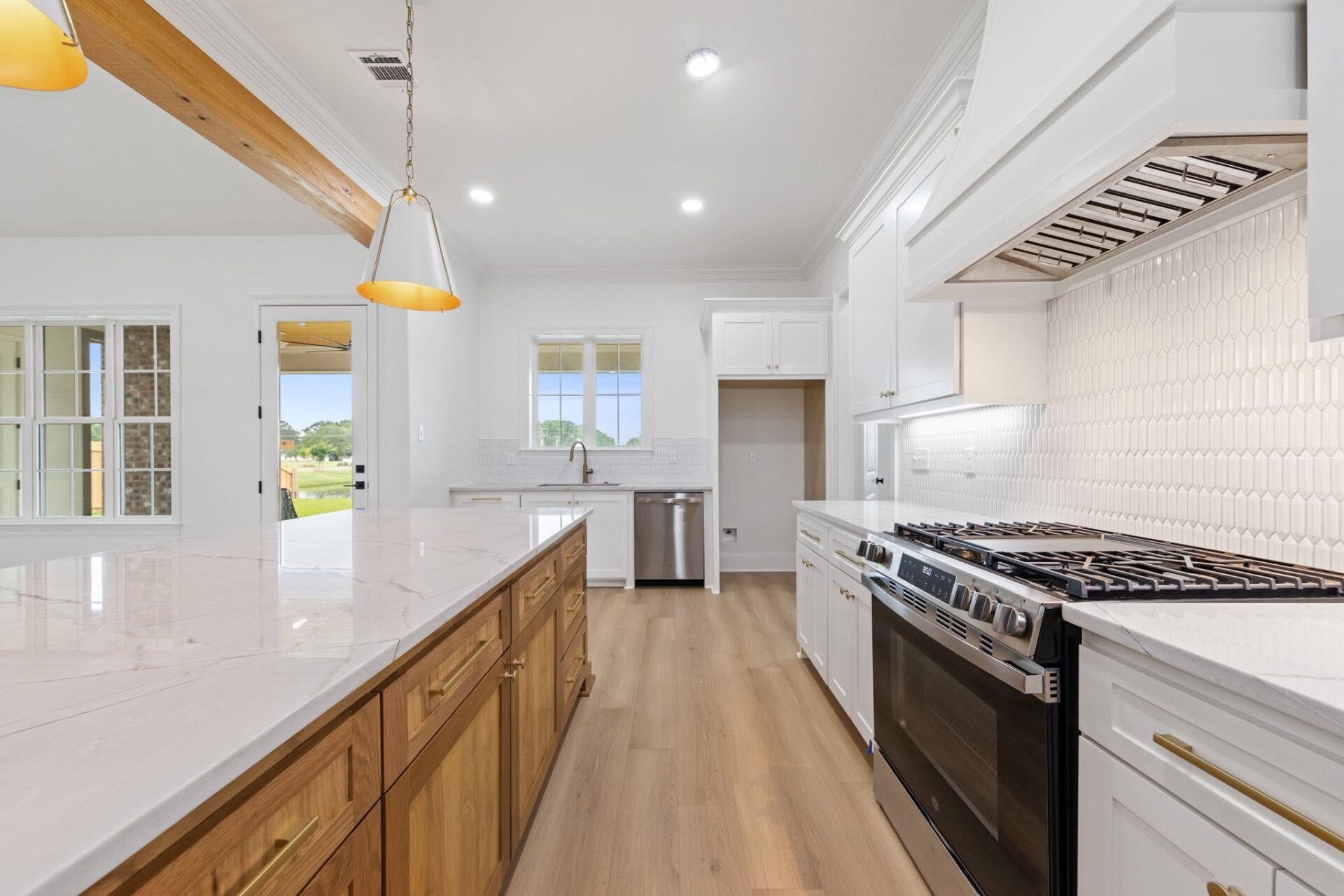 Modern kitchen with white cabinets, a large island with wood drawers, marble countertops, stainless steel appliances, and light wood flooring; pendant lights hang over the island.