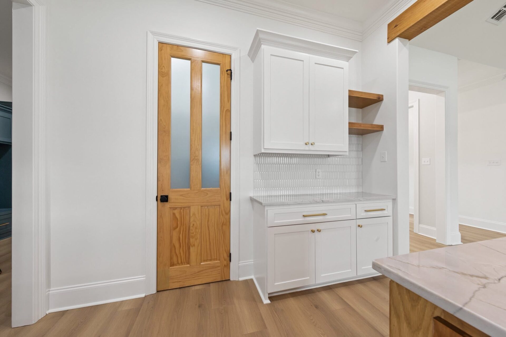 A kitchen area with white cabinets, brass handles, two wooden open shelves, a white tile backsplash, a light wood pantry door with frosted glass panels, and light wood flooring.