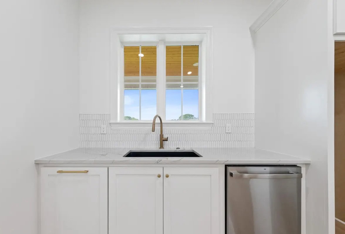 A small, modern kitchen area with white cabinets, a black sink with a gold faucet, a white tile backsplash, and a small stainless steel dishwasher under the counter. A window is above the sink.