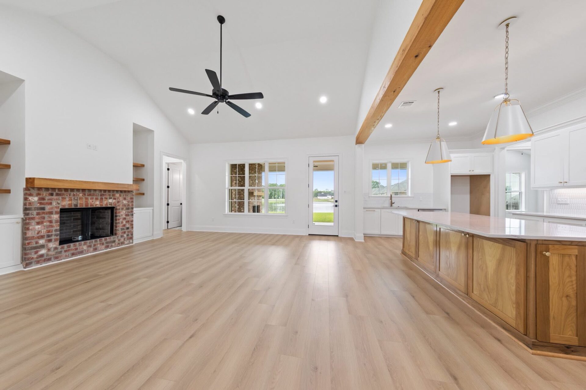 Spacious open-concept living area with light wood floors, a brick fireplace, ceiling fan, large windows, and a kitchen with a wooden island and pendant lighting. French doors lead to a green outdoor view.