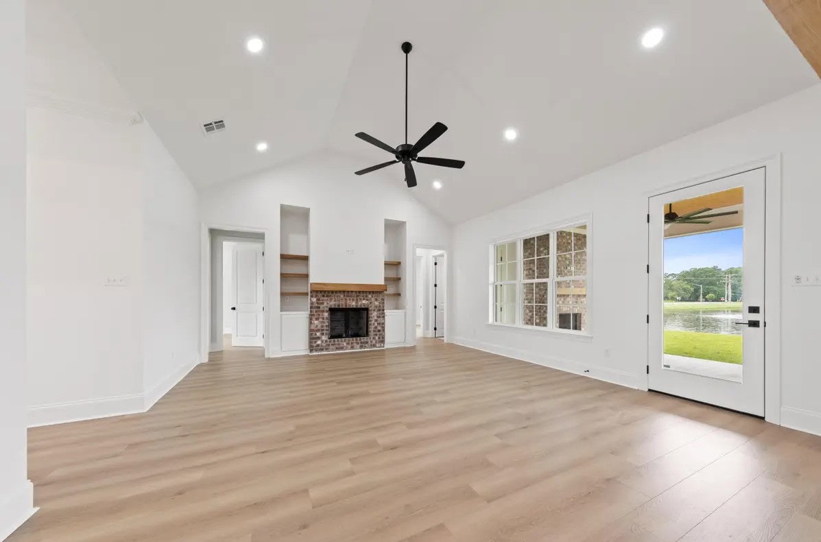 A spacious, empty living room with light wood flooring, white walls, a ceiling fan, built-in shelves around a fireplace, large windows, recessed ceiling lights, and a glass door leading outside.