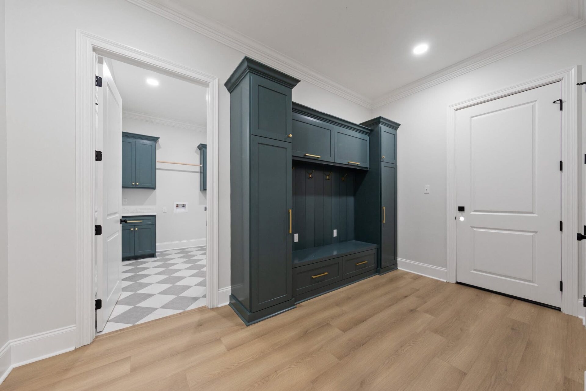 A mudroom with blue built-in cabinets, coat hooks, and drawers, adjacent to a laundry room with checkered tile flooring. The mudroom has light wood flooring and white walls with white trim.