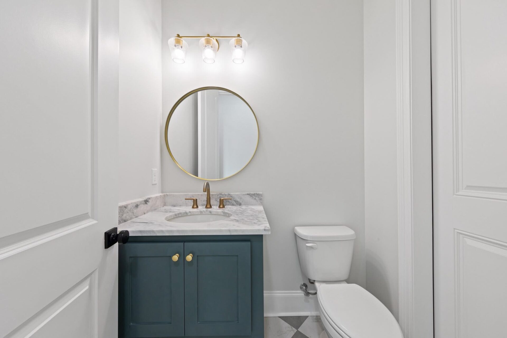 A small bathroom with a white toilet, a blue vanity with gold handles, a marble countertop, a round mirror with a gold frame, and a three-light fixture above the mirror. The walls are white.
