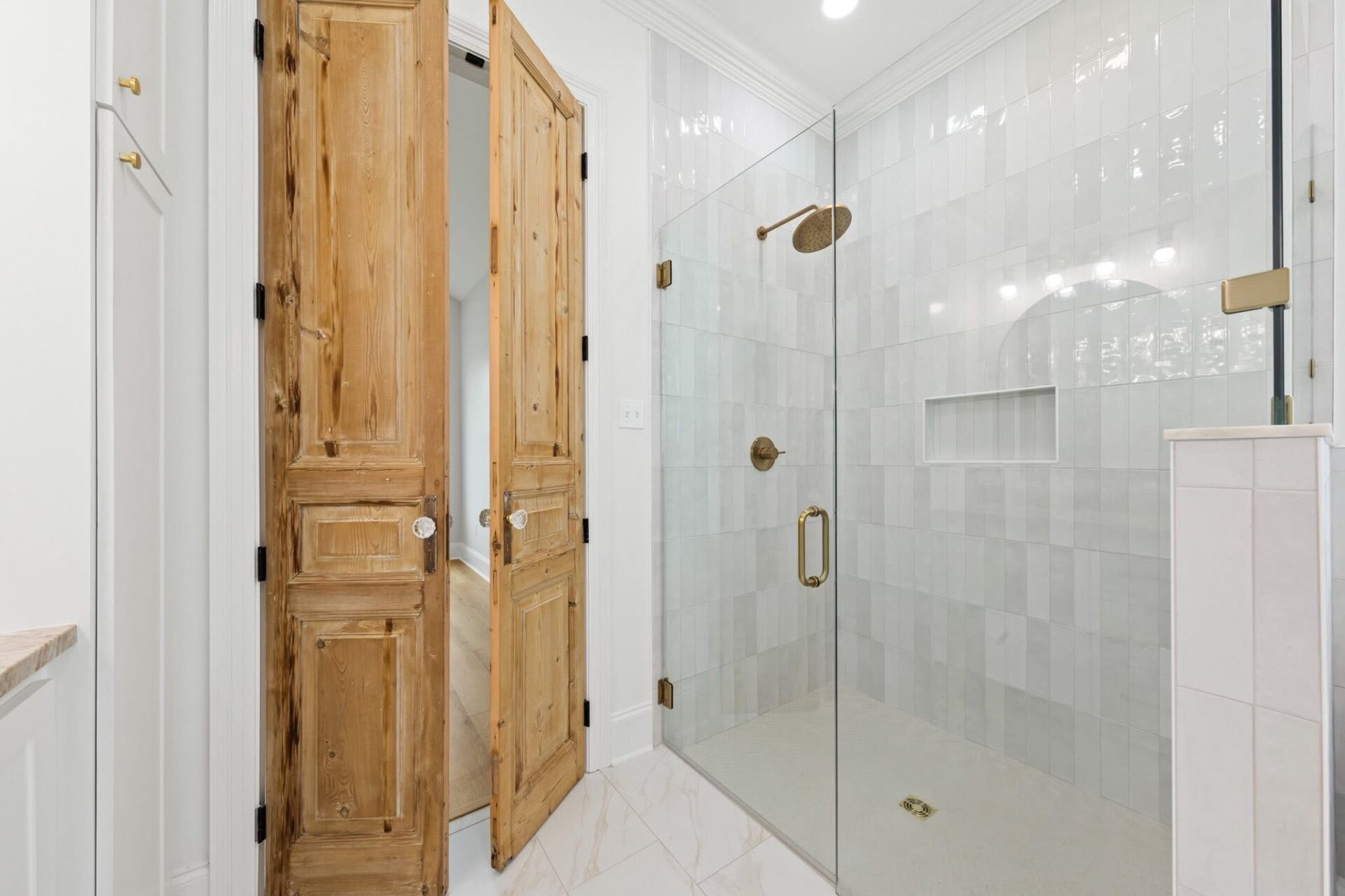 A bathroom with a large glass-enclosed shower featuring a brass showerhead and handle. The shower has white tile walls and a built-in niche. Double wooden doors and marble floor tiles are also visible.