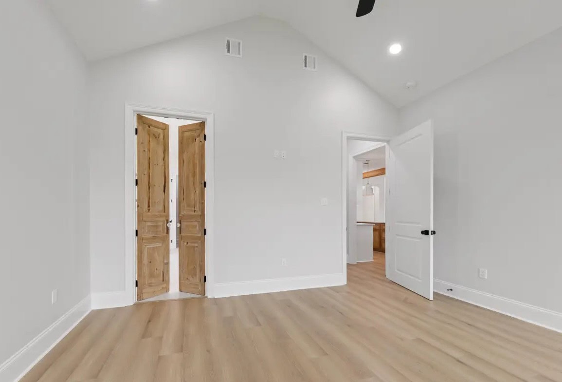 An empty room with light wood flooring, white walls, a vaulted ceiling, a set of wooden double doors, and an open white door leading to another area with visible cabinetry.