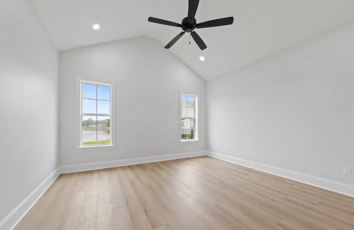 Empty room with light wood floors, white walls, two tall windows, a vaulted ceiling, recessed lighting, and a black ceiling fan. Daylight is visible through the windows.
