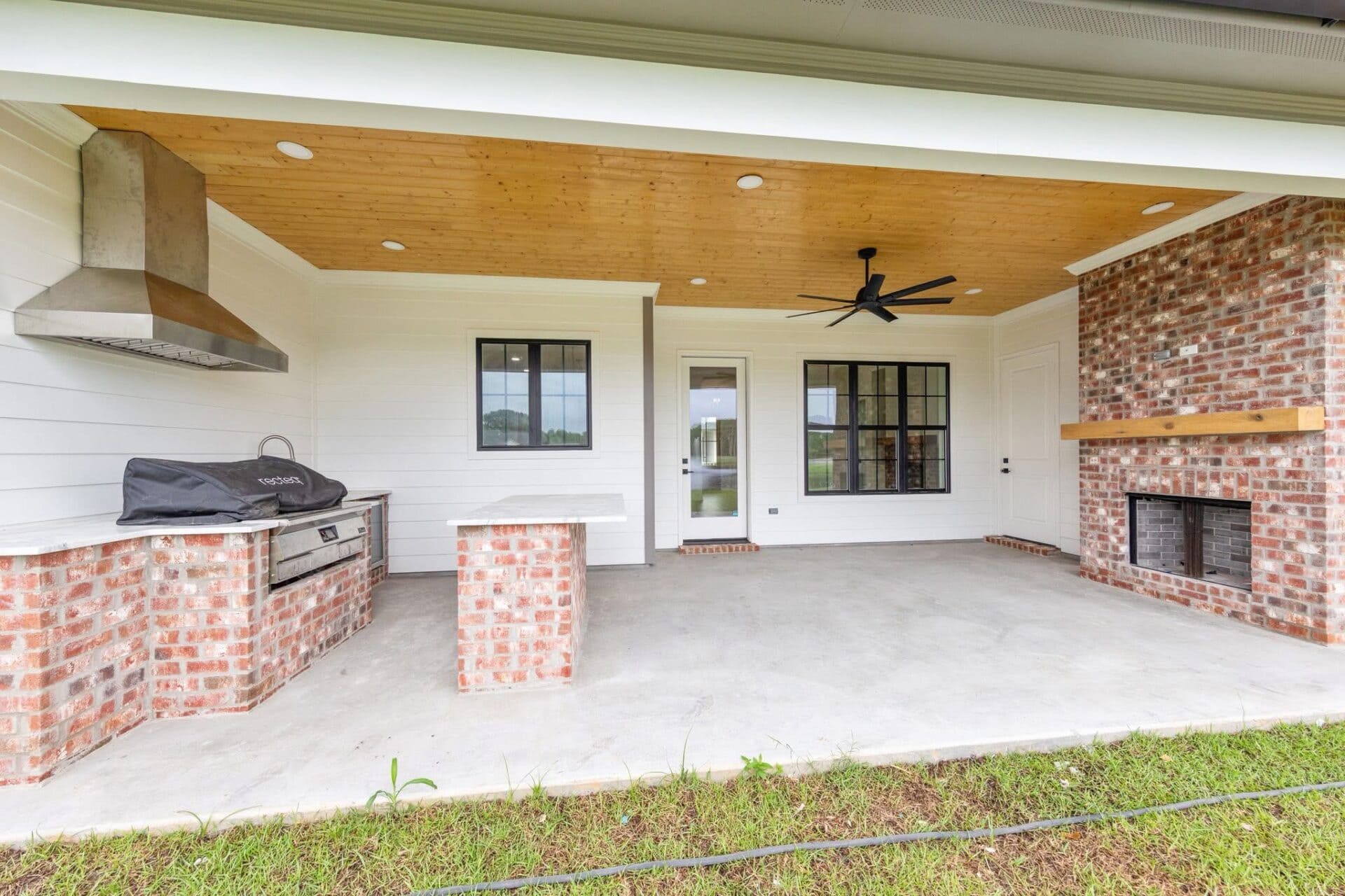 Covered outdoor patio with brick grill and oven area on the left, a ceiling fan, brick fireplace on the right, white shiplap walls, concrete floor, and glass door and windows facing the backyard.