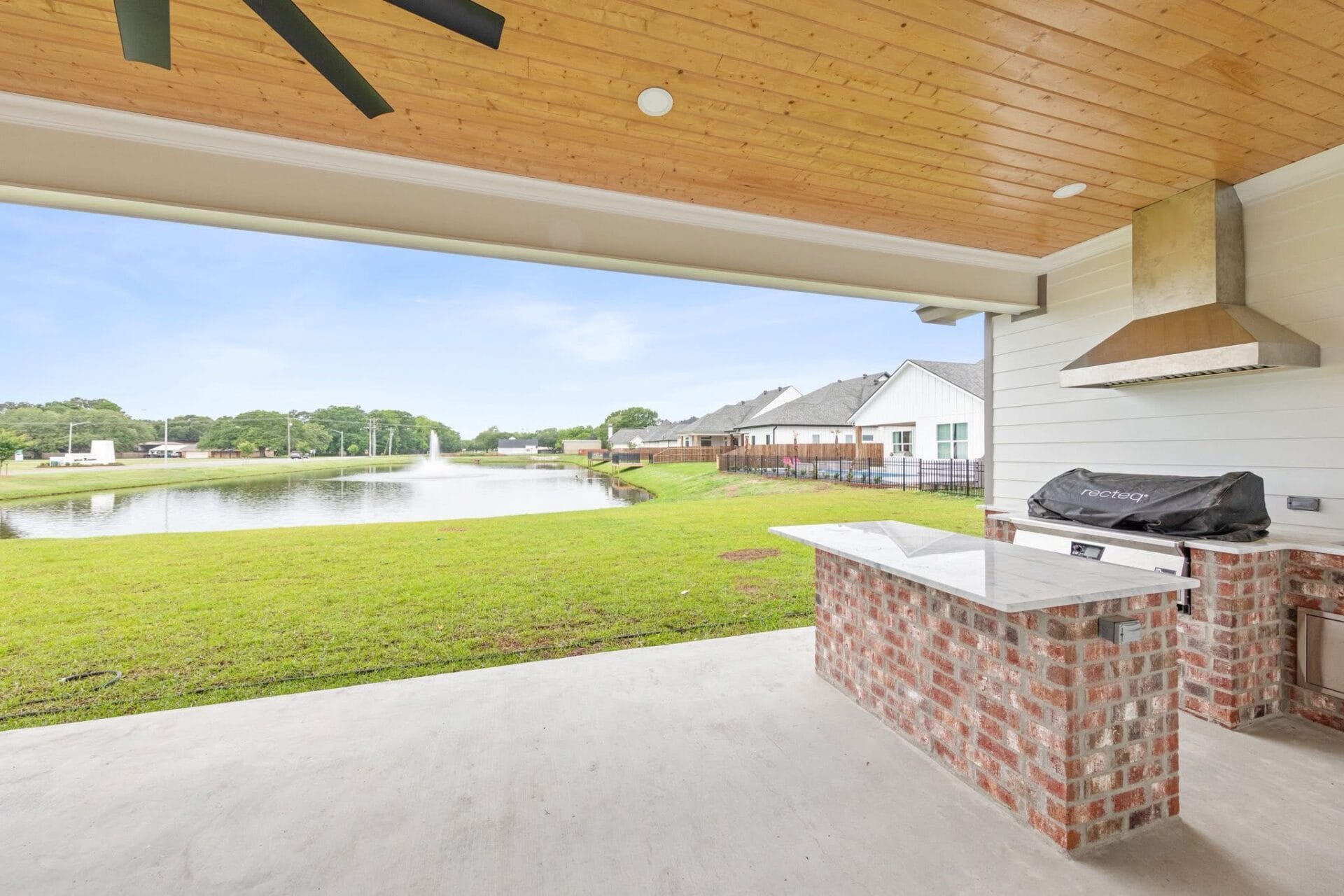 Covered patio with brick outdoor kitchen and grill, marble countertop, ceiling fan, and view of a grassy yard leading to a lake with a fountain. Houses line the opposite side of the lake under a clear sky.