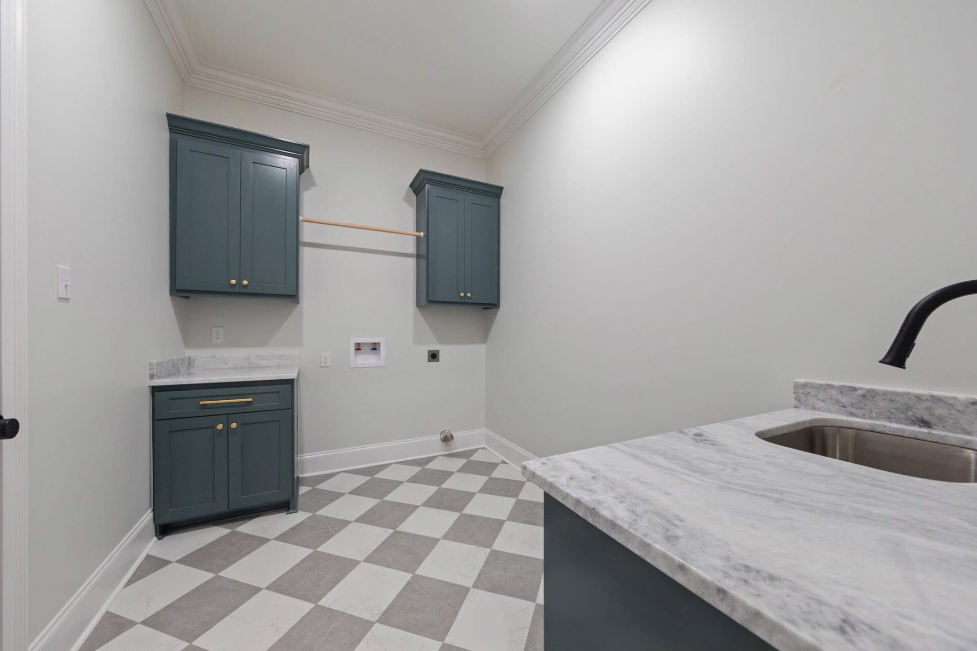 A laundry room with gray and white checkered floor, blue-green cabinets with gold handles, an empty countertop with a sink, and wall outlets for a washer and dryer. The walls are light-colored.