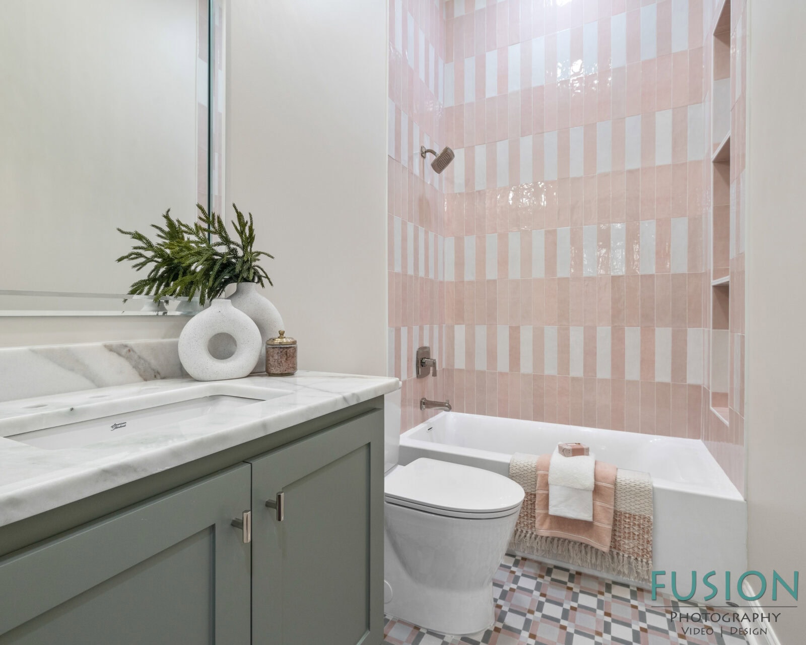 Modern bathroom with sage green vanity, marble countertop, rectangular mirror, white toilet, and a bathtub with pink and white vertical striped tile. Towels and a plant decor are neatly arranged. Fusion Photography watermark is visible.