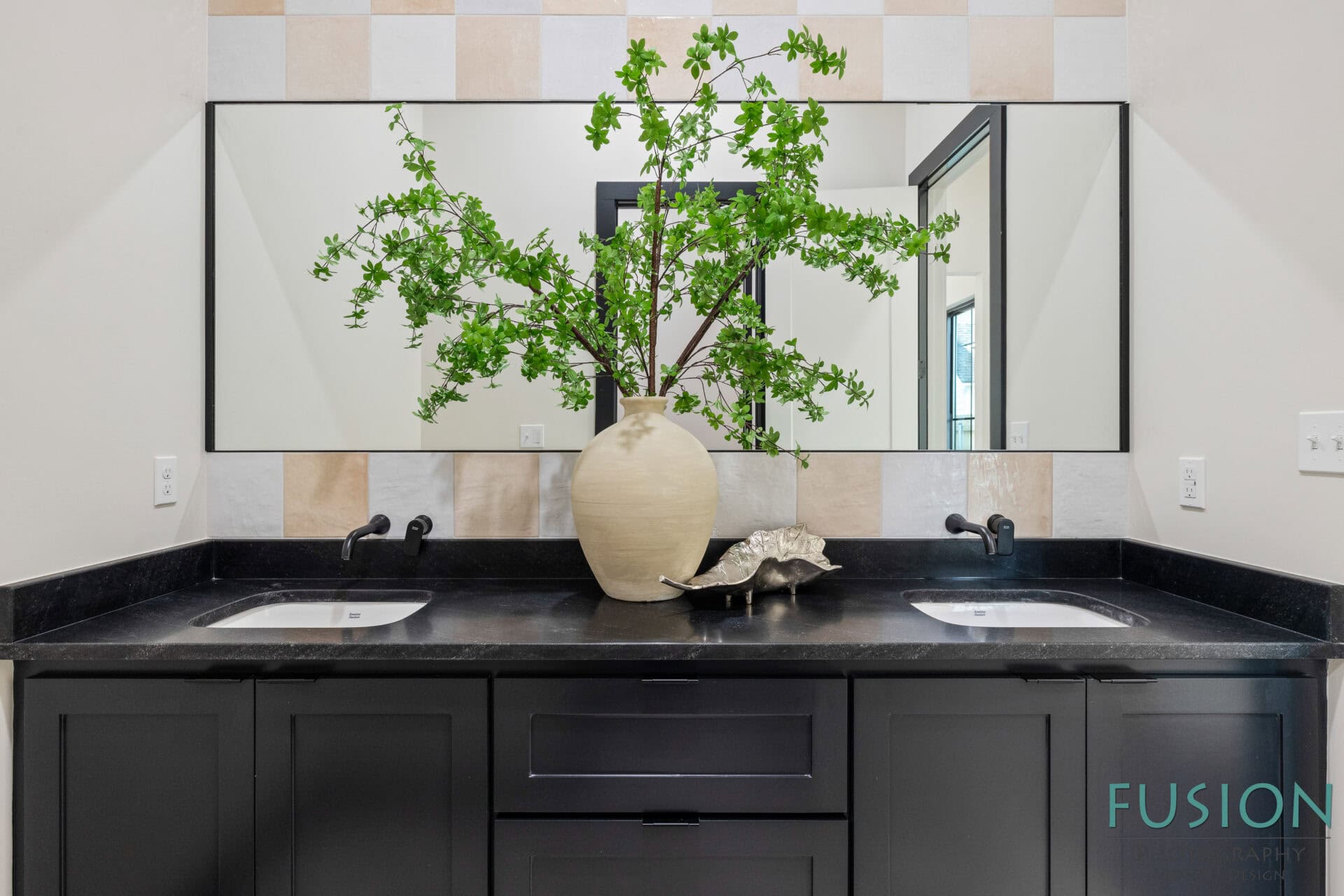 A modern bathroom vanity with black cabinets, a black countertop, two sinks, a large rectangular mirror, a beige vase with green branches, and a decorative shell. Light-colored tiles are on the wall behind the mirror.