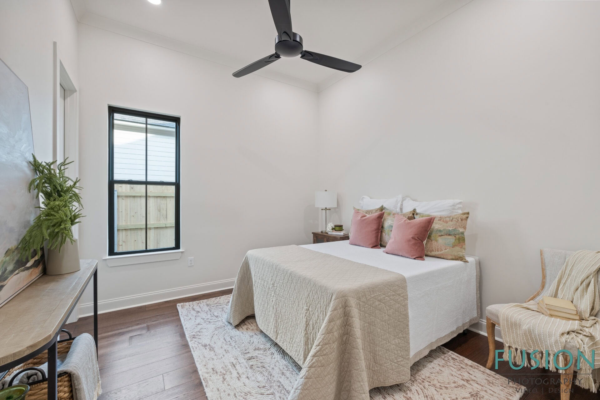 A small bedroom with a double bed, light bedding, and pink pillows. A window with blinds is on the left wall, and a ceiling fan hangs above. There is a side table with a lamp and a chair with a blanket in the corner.