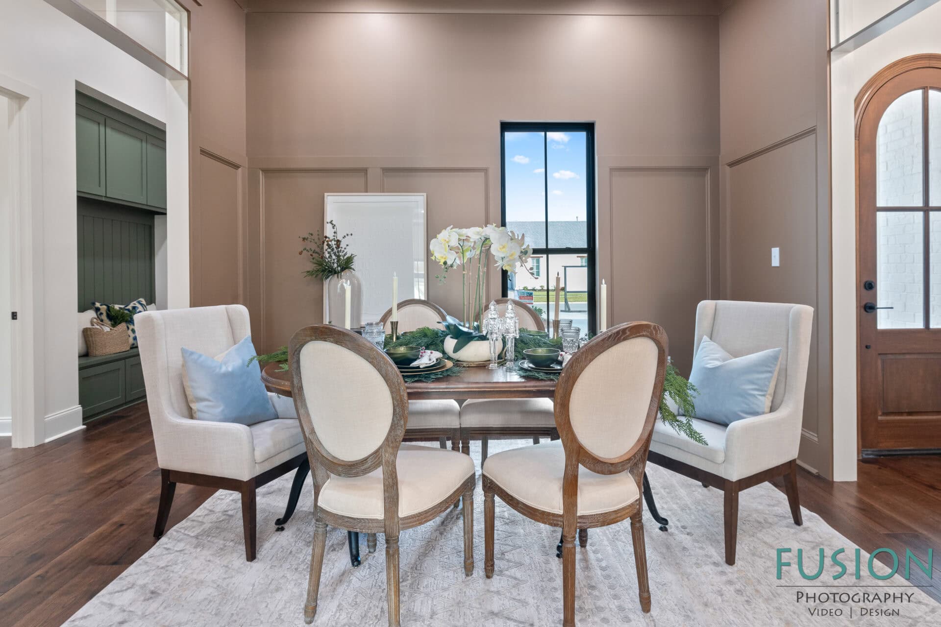 A dining room with a wooden table set for four, surrounded by beige upholstered chairs and a green accent wall on the left. A tall window lets in natural light, and the floor is dark wood with a light area rug.