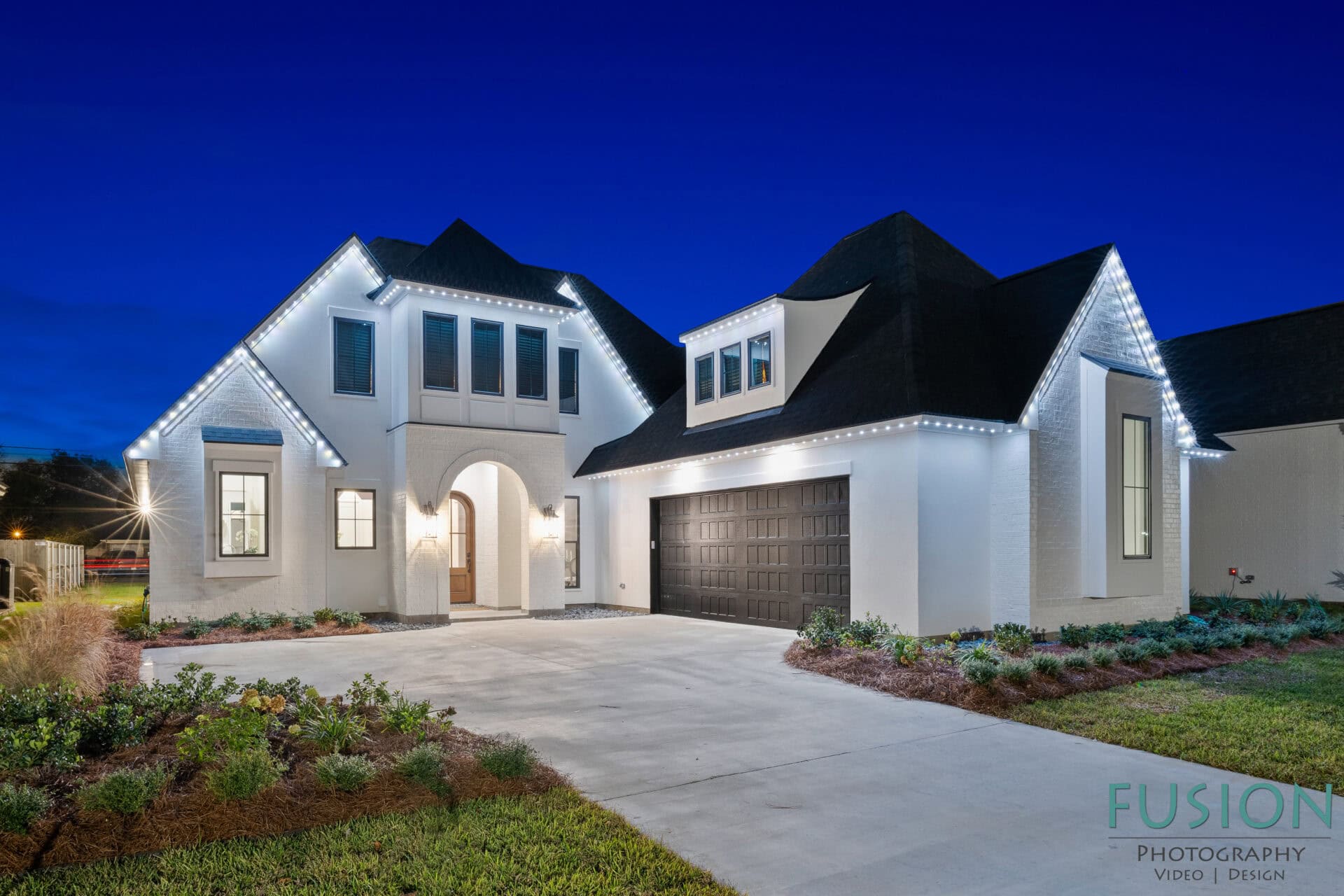 A modern white house with dark roof shingles, a two-car garage, and exterior LED lights outlining the roof, photographed at dusk with a clear blue sky. Manicured landscaping lines the driveway and entrance.