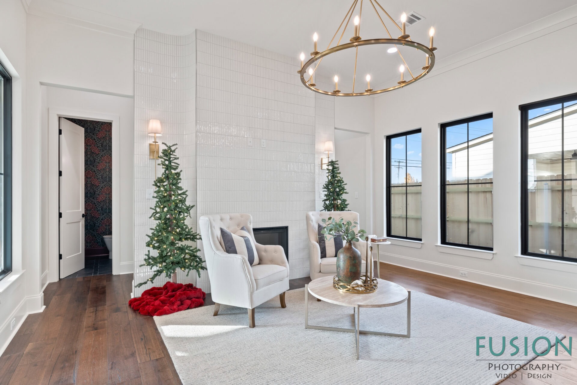 A modern living room with two tufted armchairs, a round coffee table, two artificial Christmas trees by a white-tiled fireplace, a brass chandelier, and three large windows with black frames.