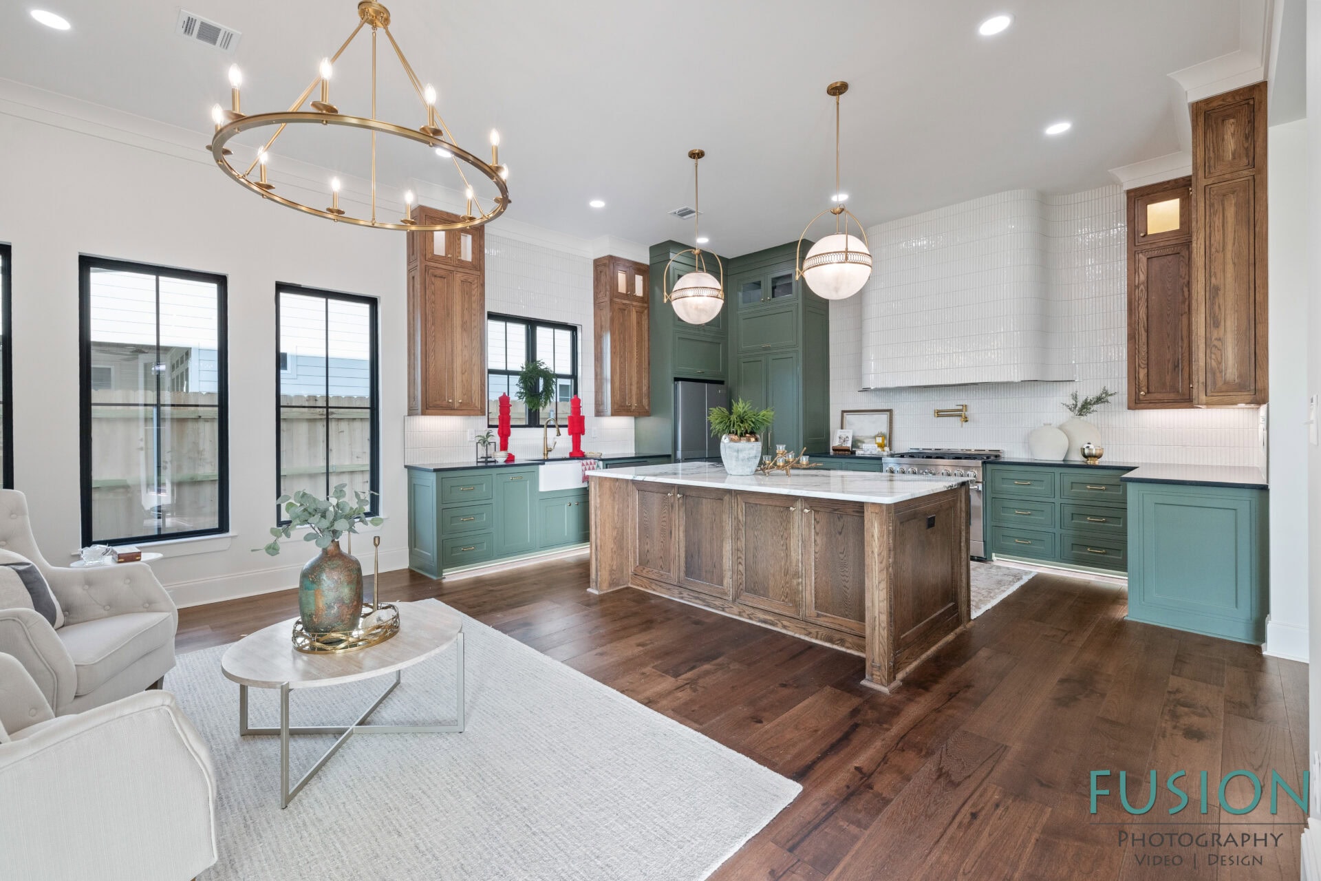 A modern kitchen with green cabinets, a large wooden island, white marble countertops, wooden flooring, gold light fixtures, and a small sitting area with a white couch and round coffee table. Fusion Photography watermark is visible.