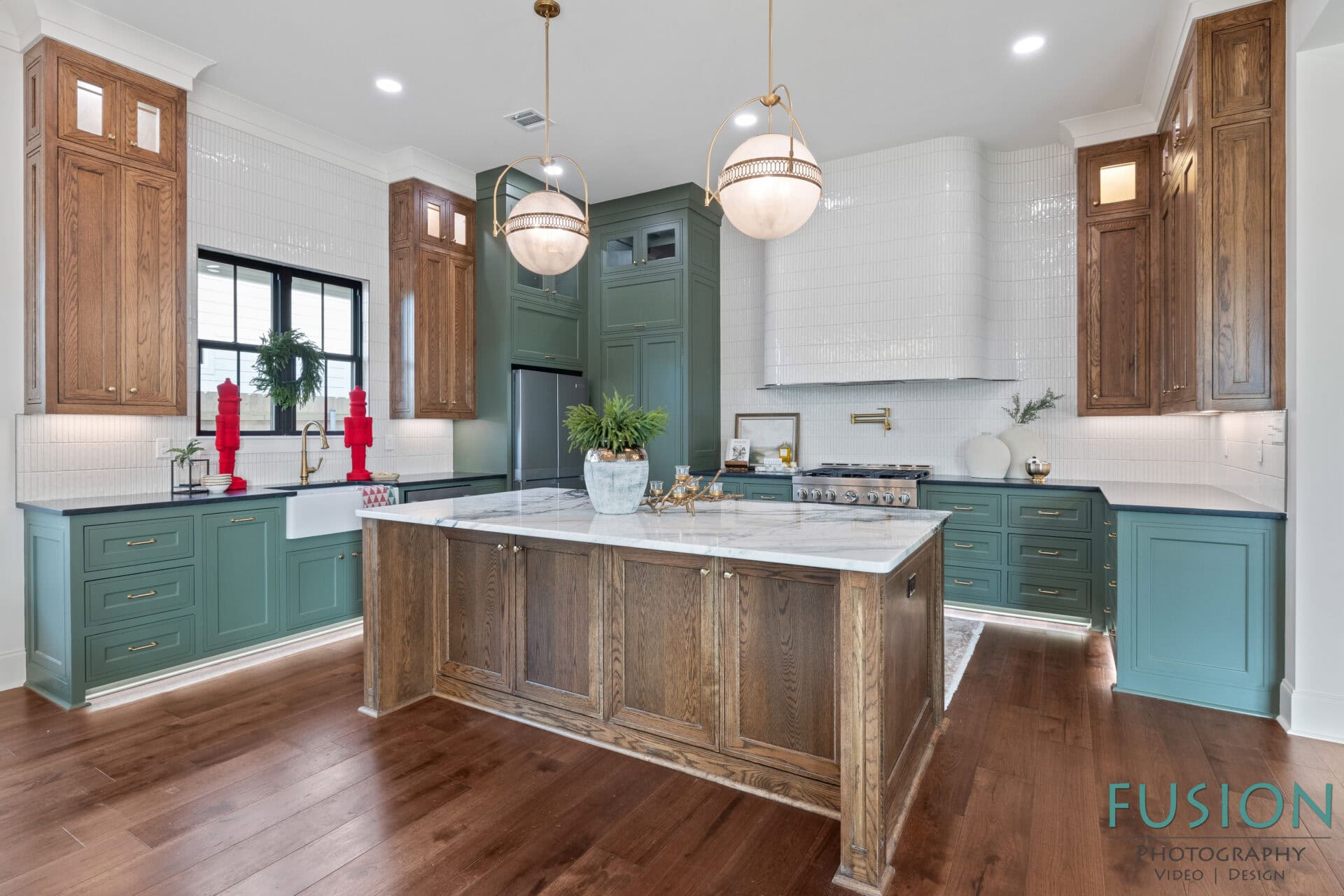 A spacious kitchen features green lower cabinets, wood accents, a marble island countertop, white tiled walls, stainless steel appliances, pendant lights, and decorative red candlesticks near a window.